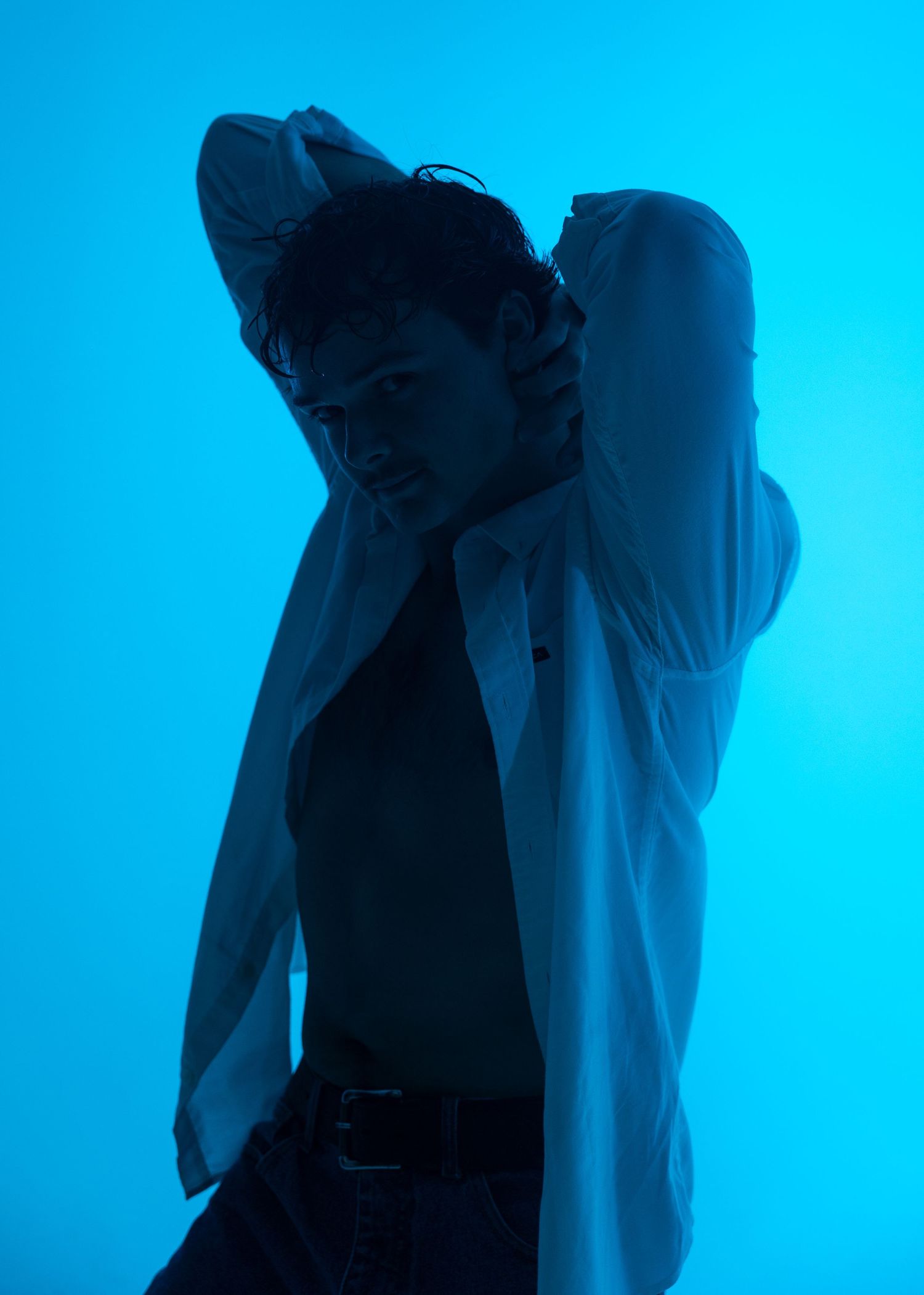 A young man poses dramatically in a white shirt under moody blue lighting, hands raised behind his head.
