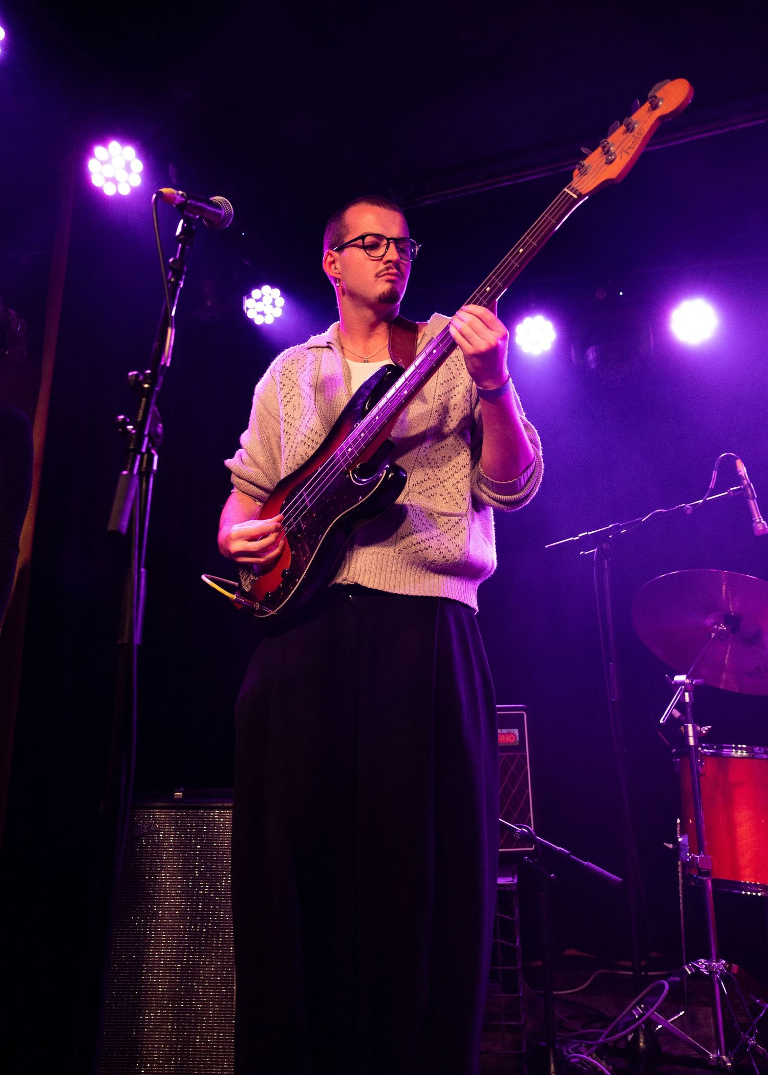Musician playing bass guitar on stage under purple lights, wearing glasses and a white sweater with a black skirt.