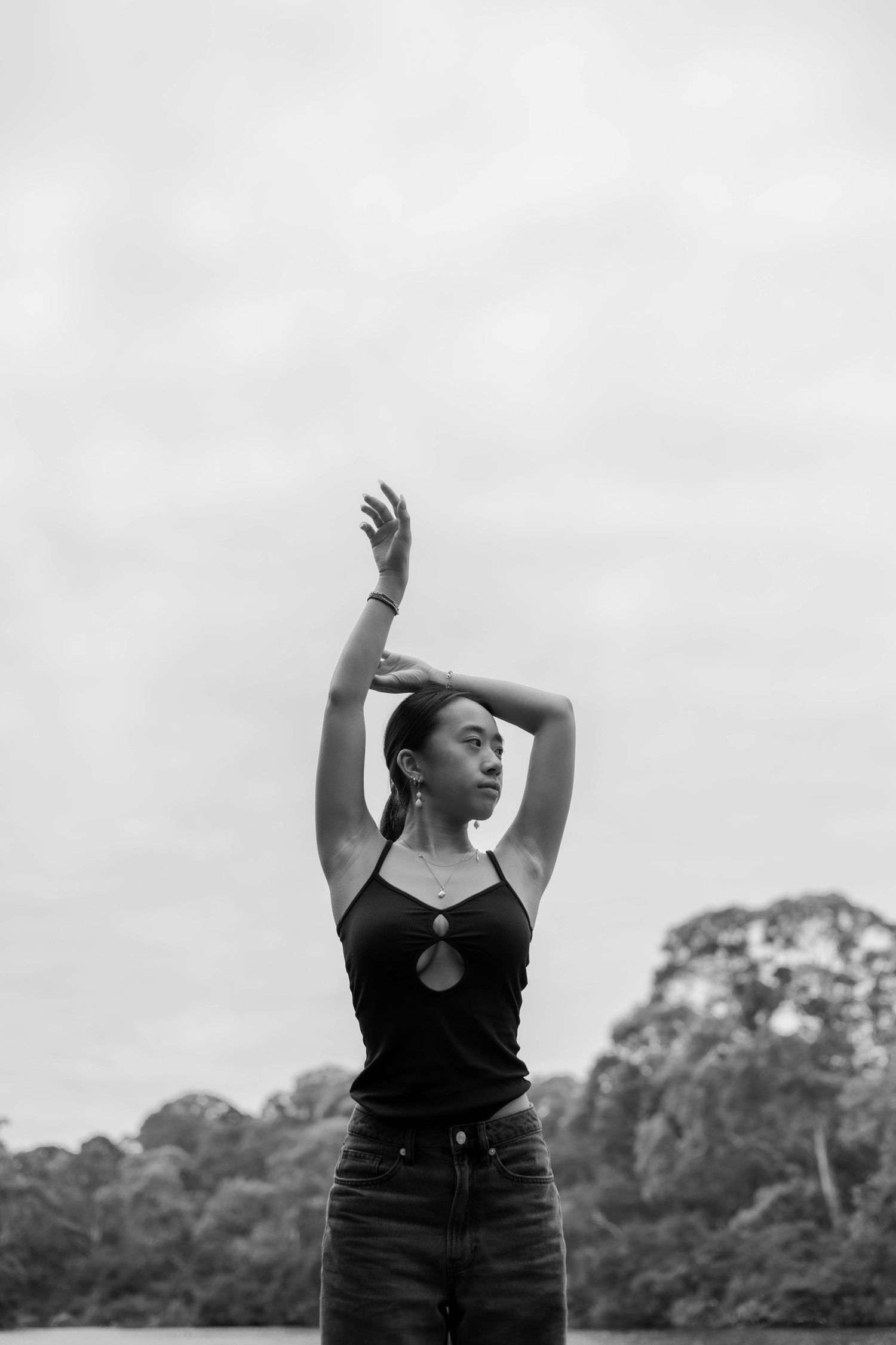 Black and white photo of a young woman posing outdoors with arms raised above her head against a cloudy sky.