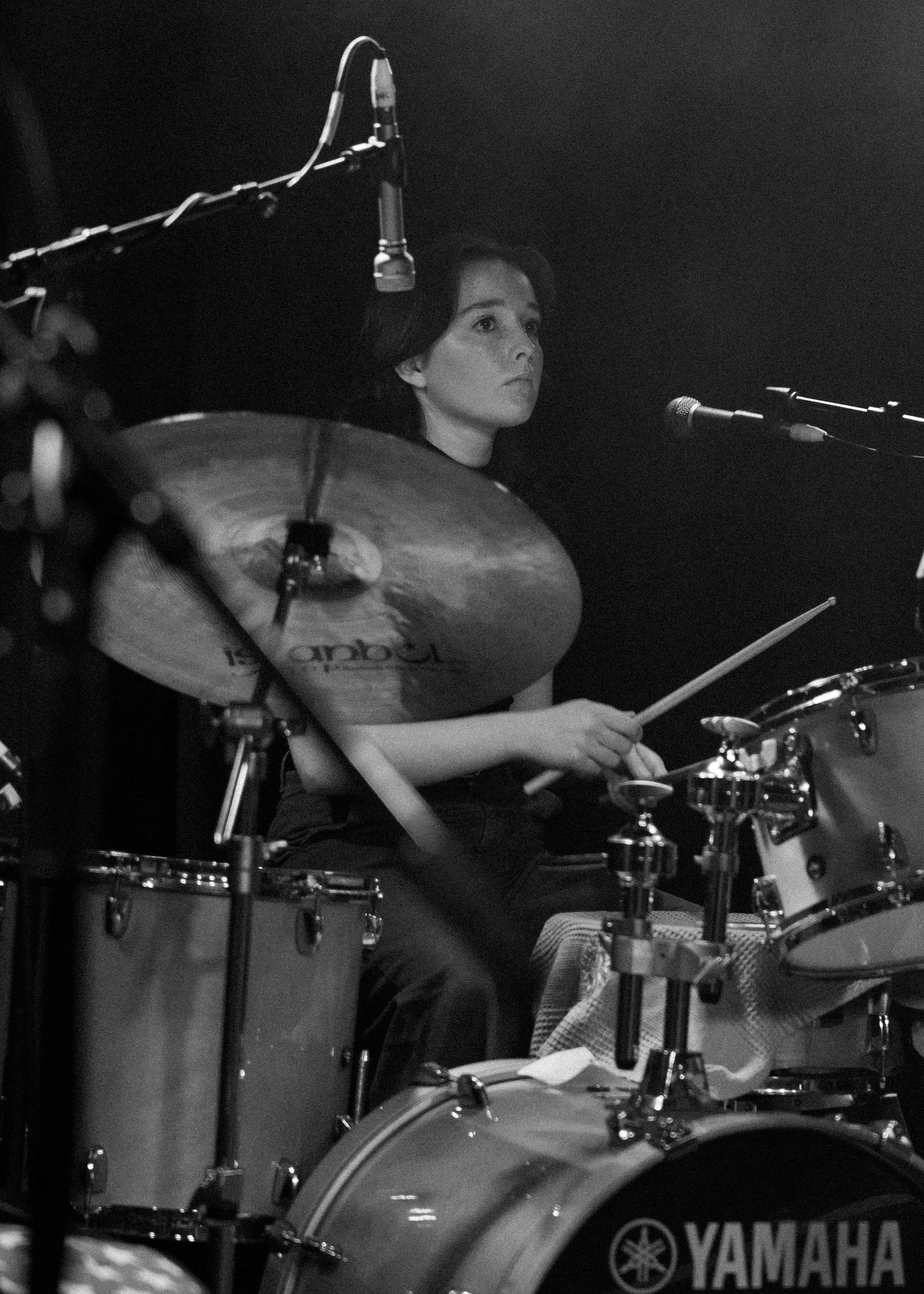 Black and white photo of a young woman playing Yamaha drums on stage with cymbals and microphones visible.