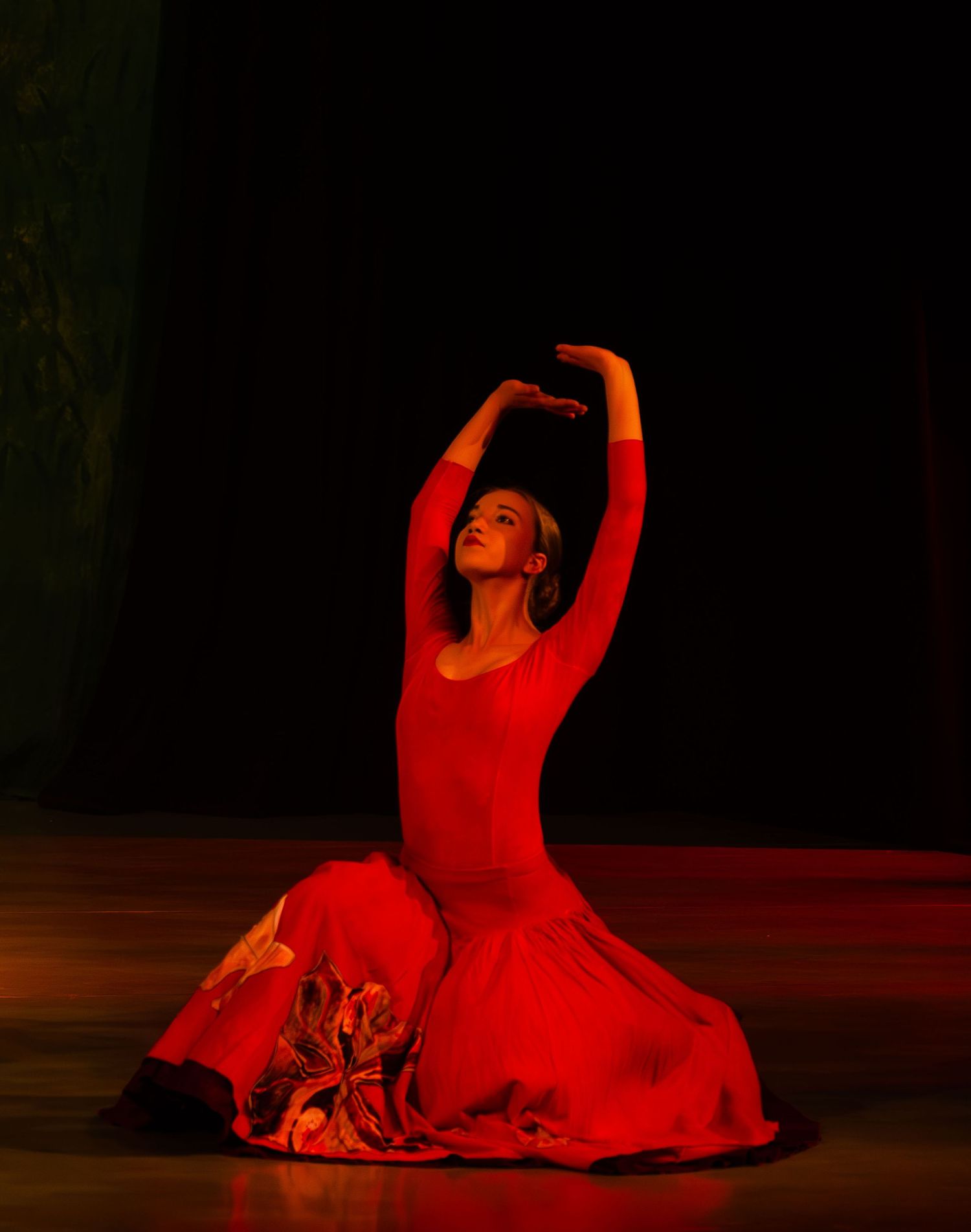 A dancer in flowing red costume performs an expressive floor movement sequence in dramatic lighting.