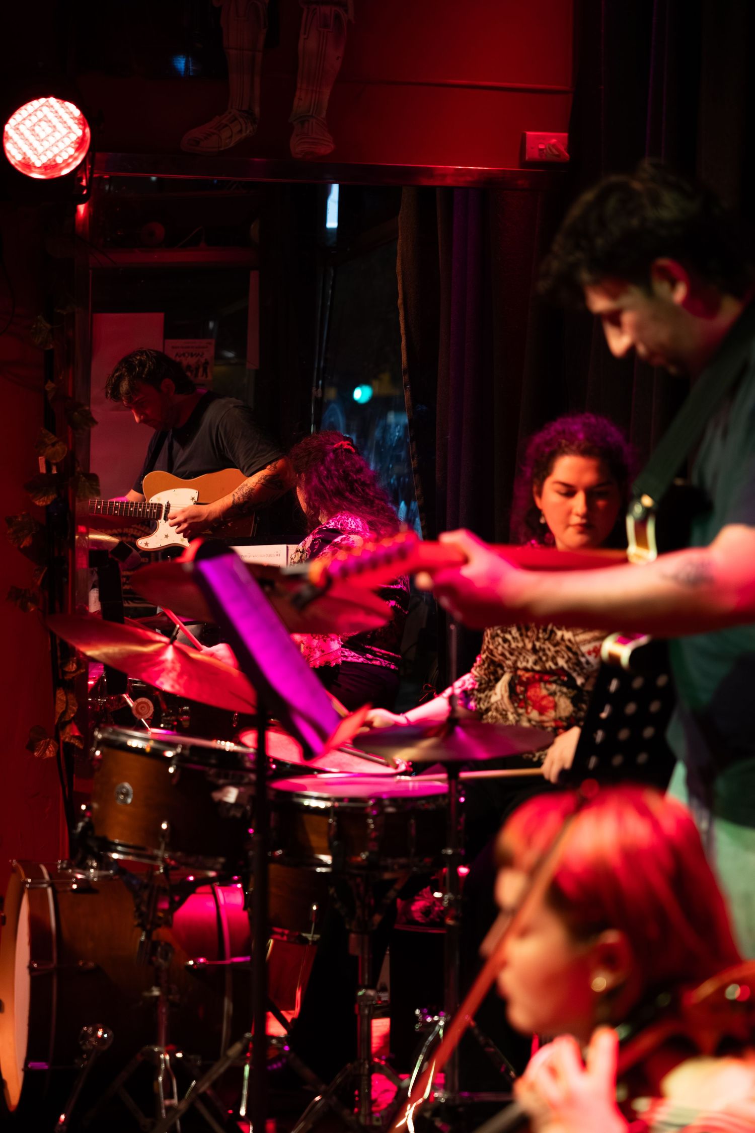 Musicians perform in a dimly lit venue with red lighting and drum kit visible in the foreground.