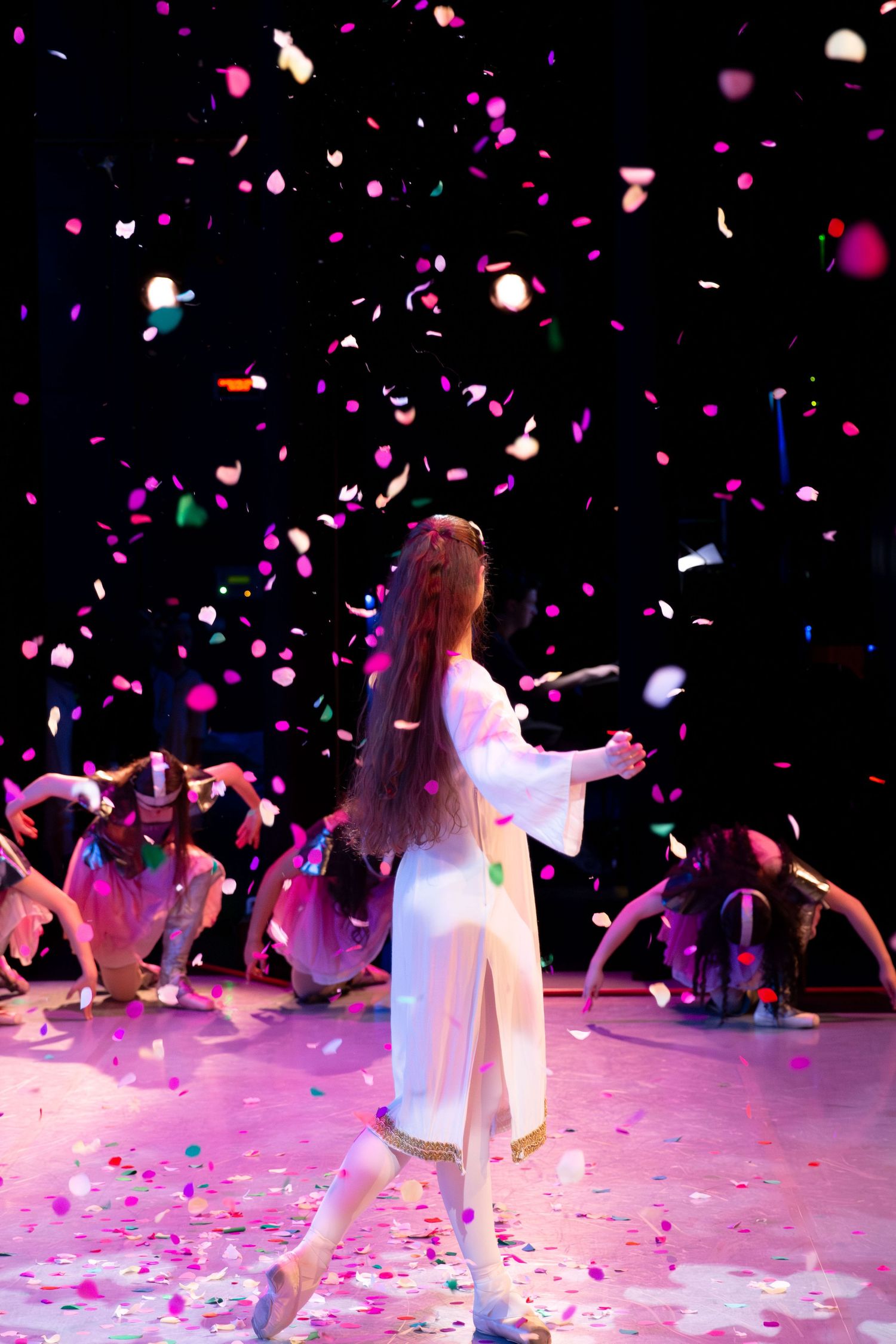 A dancer in white performs on stage amid colorful falling confetti with other performers in the background.