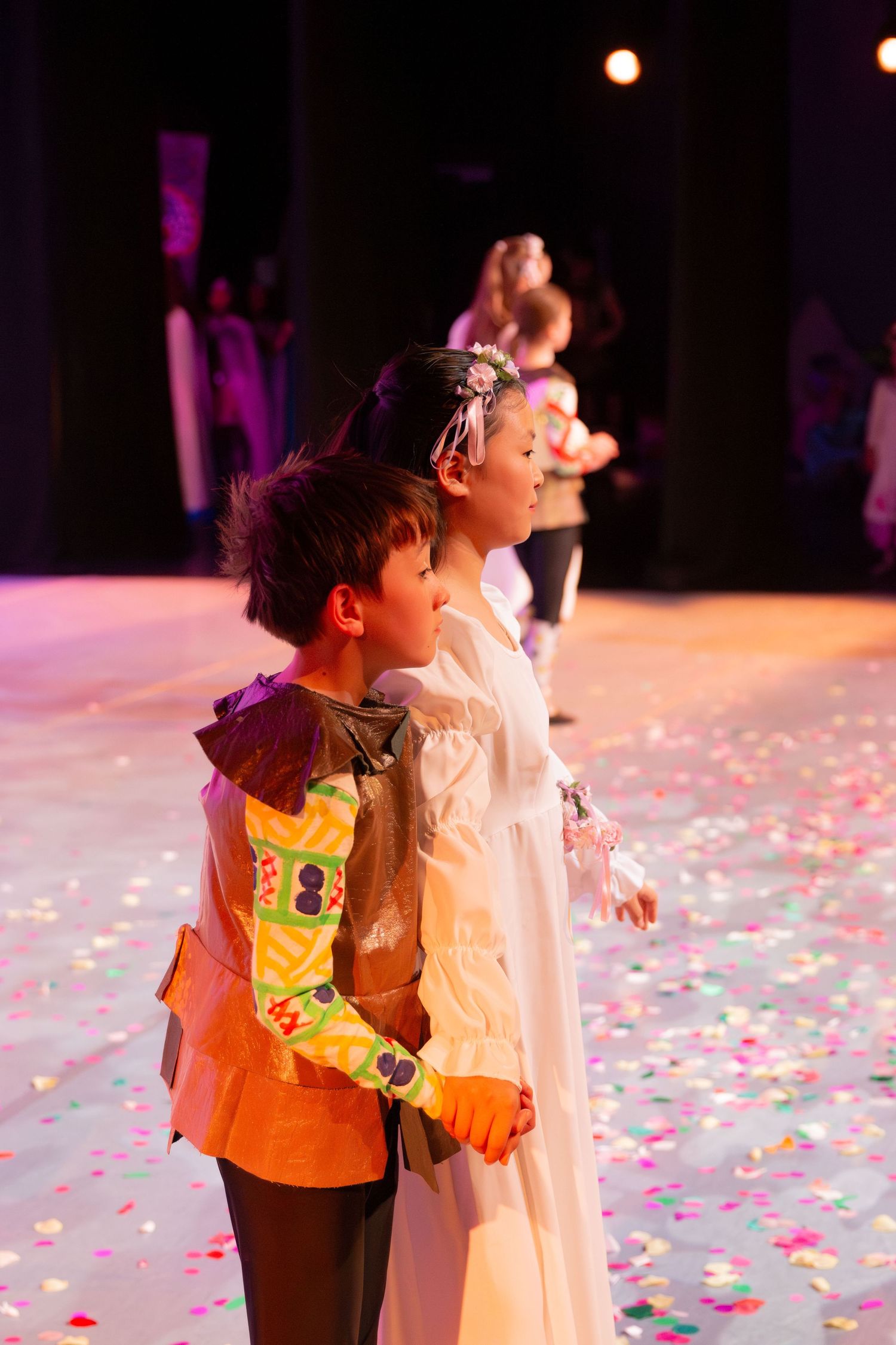 Children in costumes perform on a confetti-covered stage under colorful lights during a school theater production.