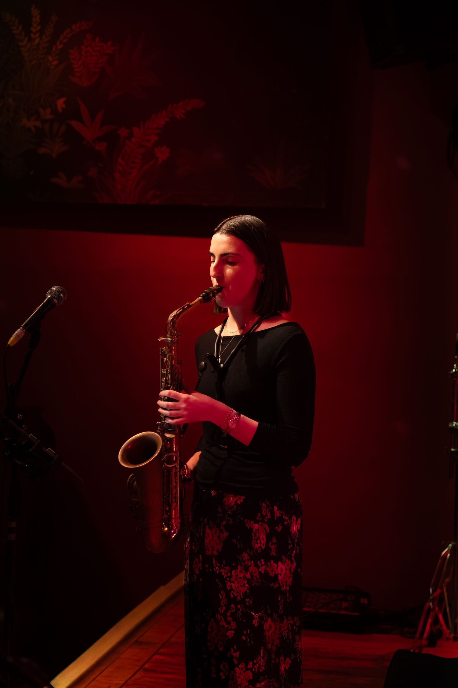 Musician plays saxophone against dark background illuminated by moody red stage lights.