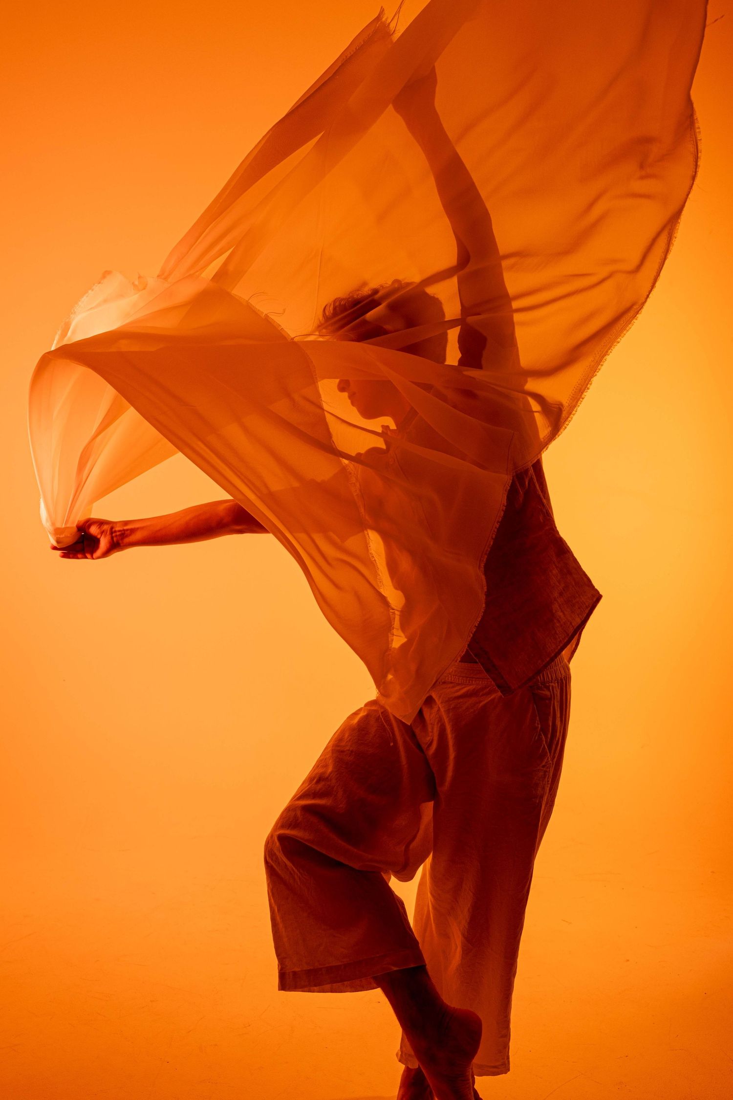 Artistic silhouette of dancer with billowing fabric creating movement against orange background.