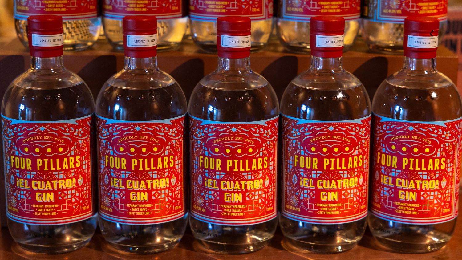 Four Pillars El Cuatro gin bottles displayed in a row on a wooden shelf with red and gold labels.