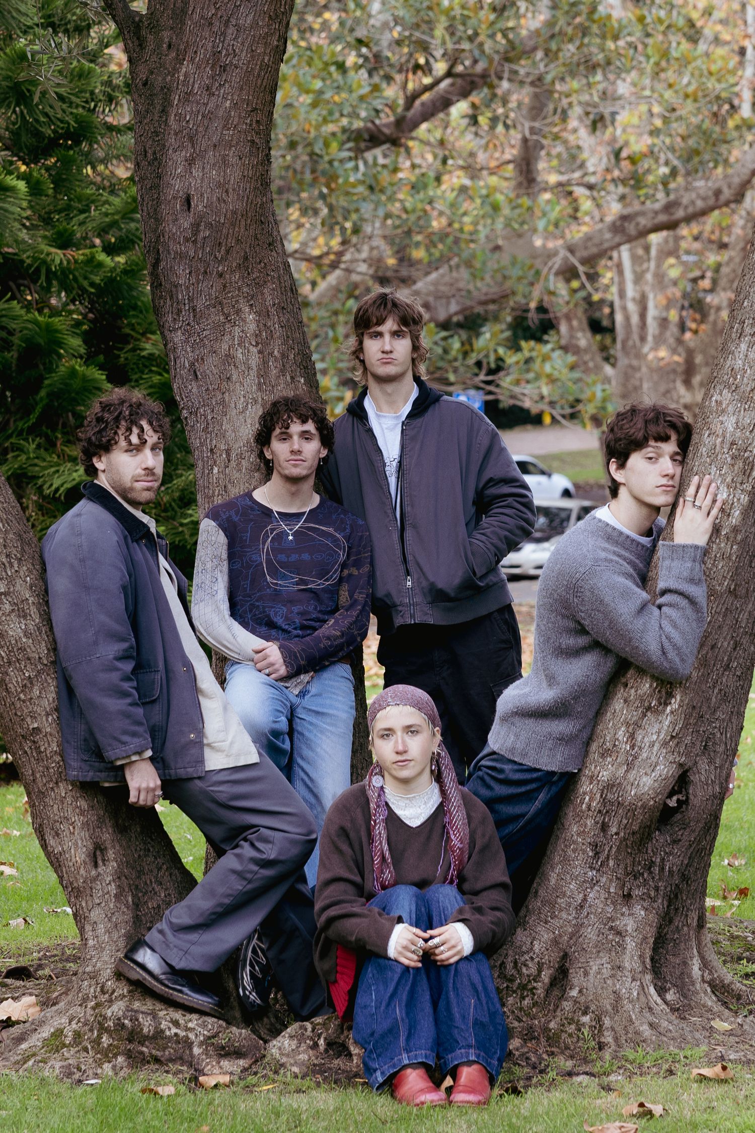 Five young people pose between large tree trunks in a lush green park.