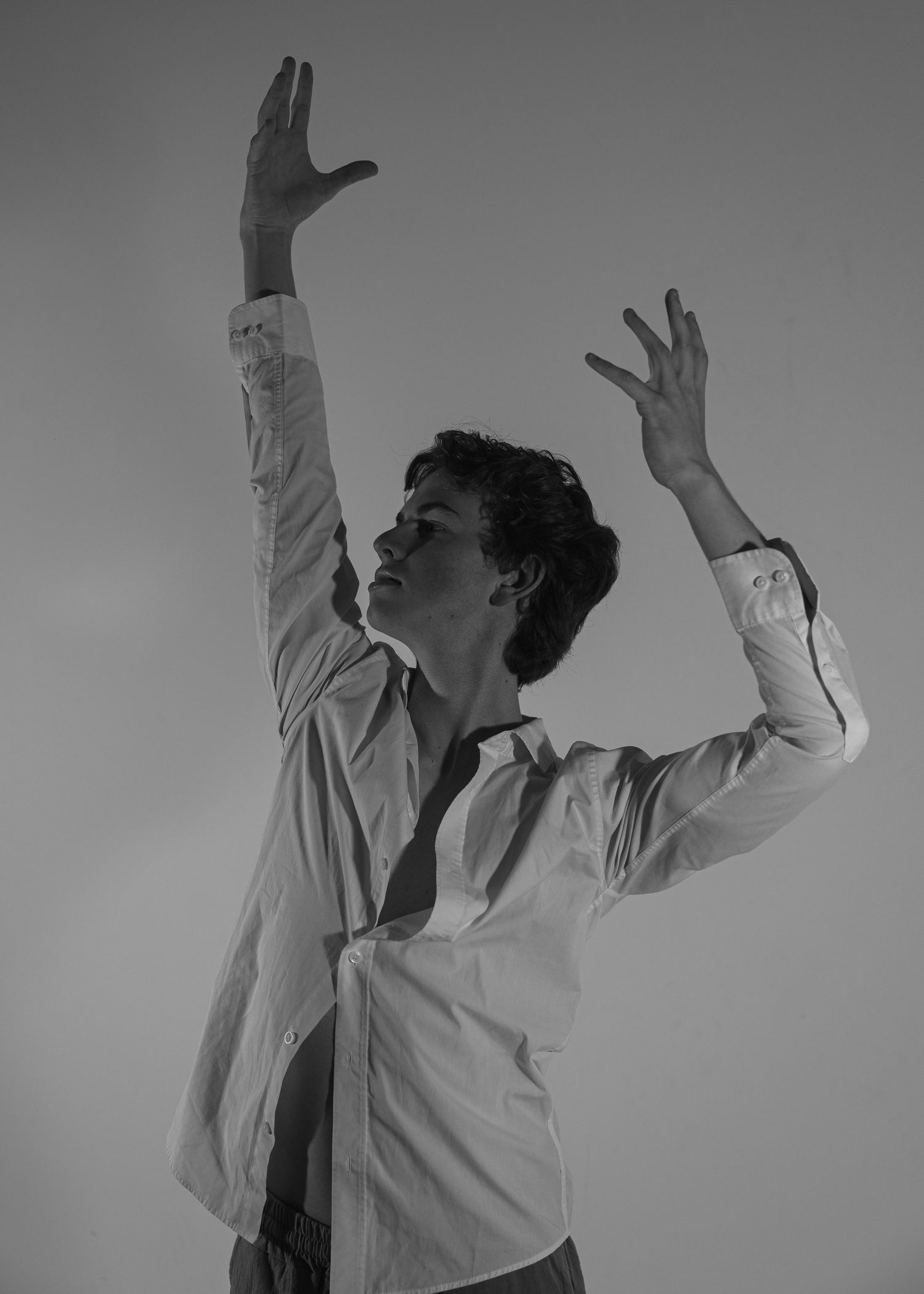 Black and white photo of a dancer in a white shirt with both arms raised gracefully overhead.