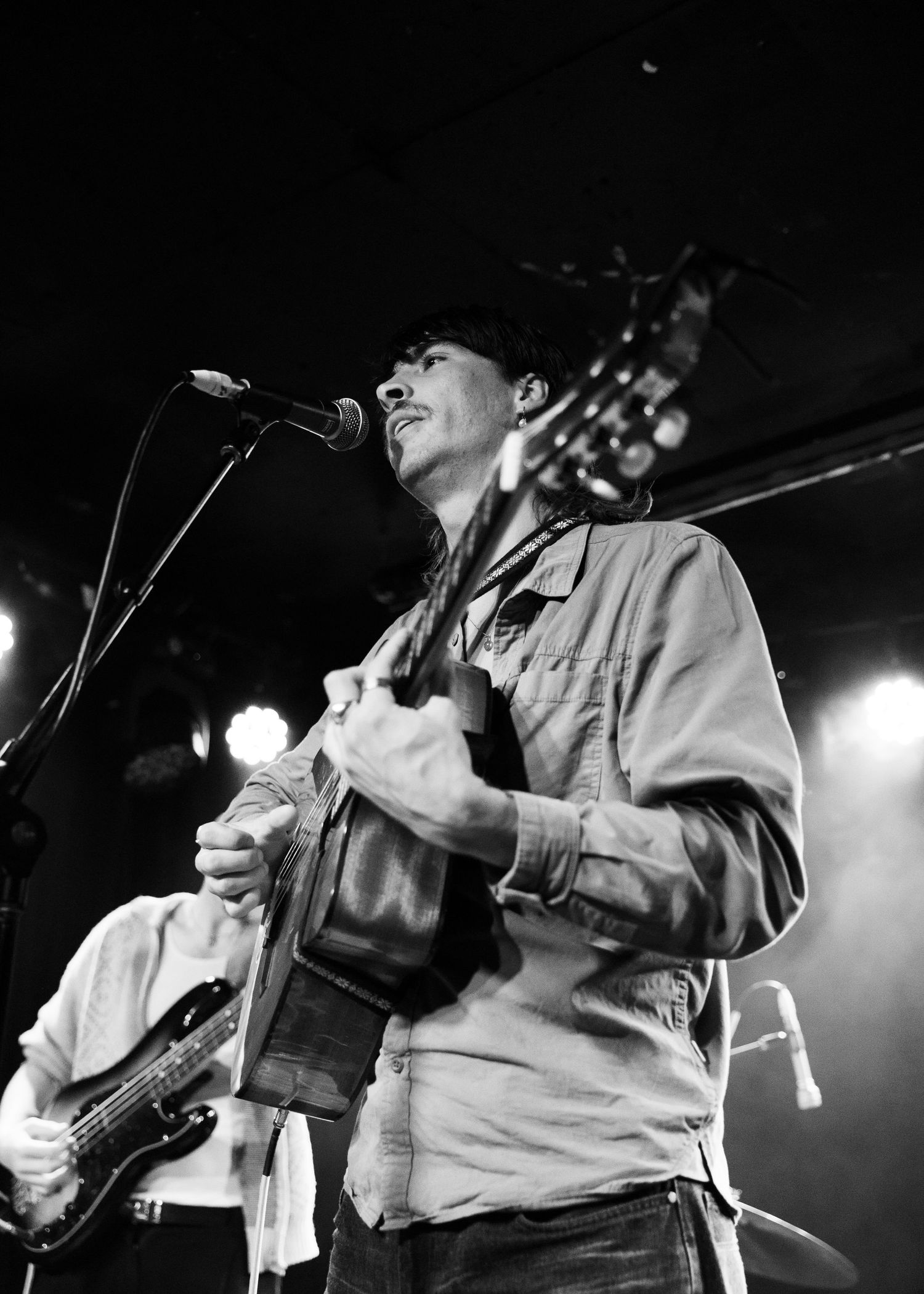 Black and white photo of a male guitarist singing into a microphone on stage during a live performance.