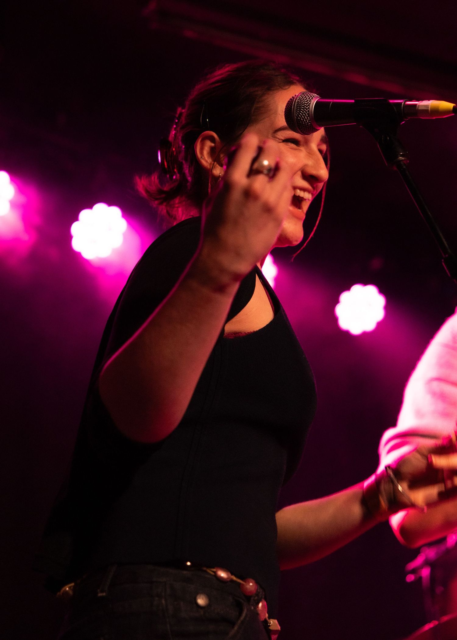 Female vocalist performing on stage with microphone, pink stage lights glowing behind her at a live concert.