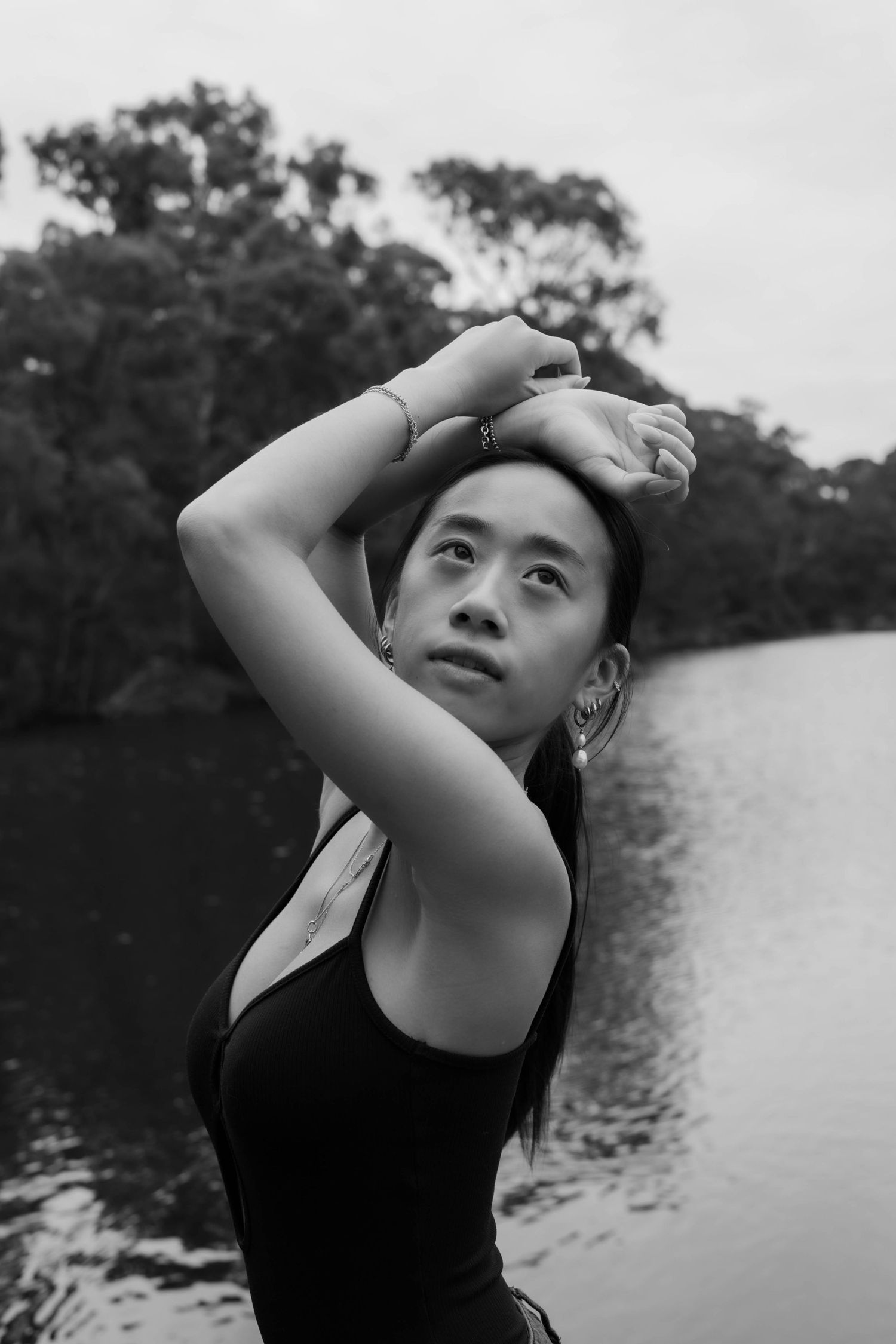 Black and white portrait of a young Asian woman posing by a lake with trees, arms raised gracefully overhead.