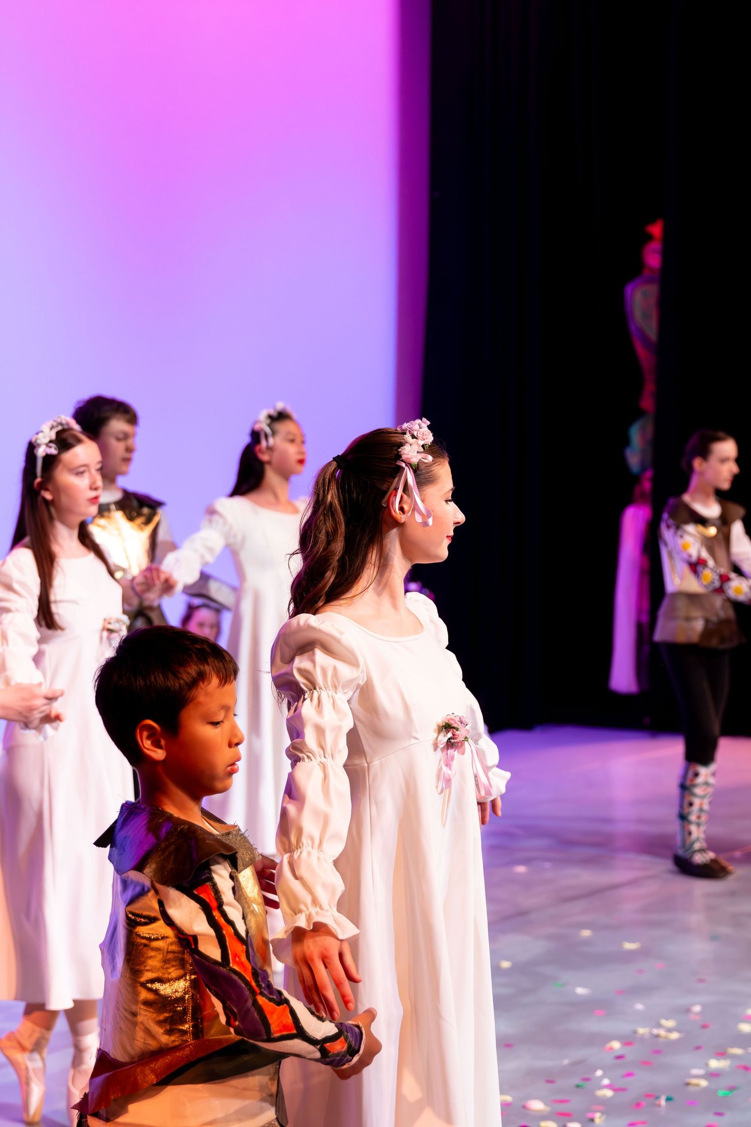 Children perform in a ballet recital on stage, wearing white and colorful costumes under pink and purple stage lighting.