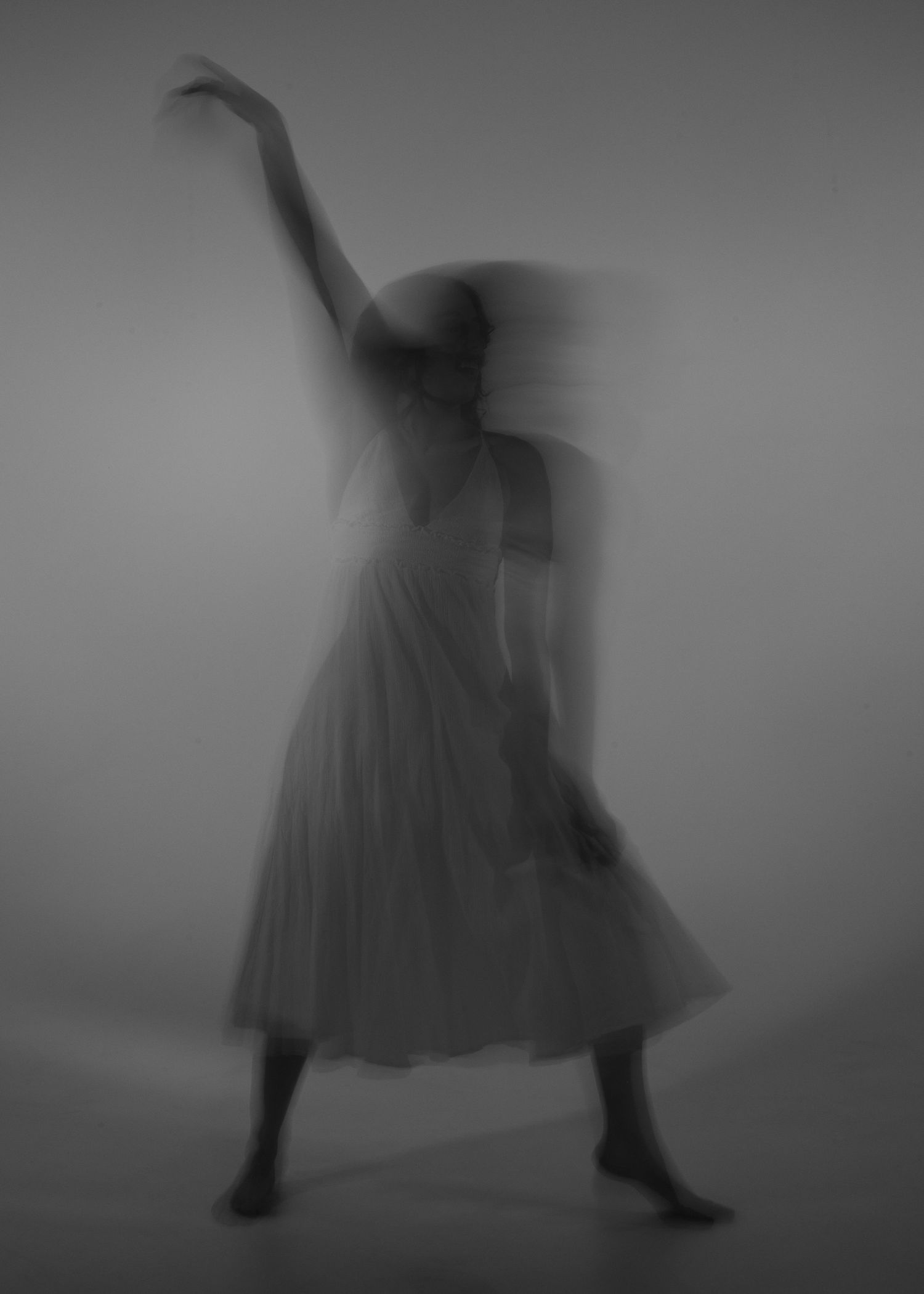 Blurred black and white long exposure photo of dancer in flowing dress with arm raised mid-movement.