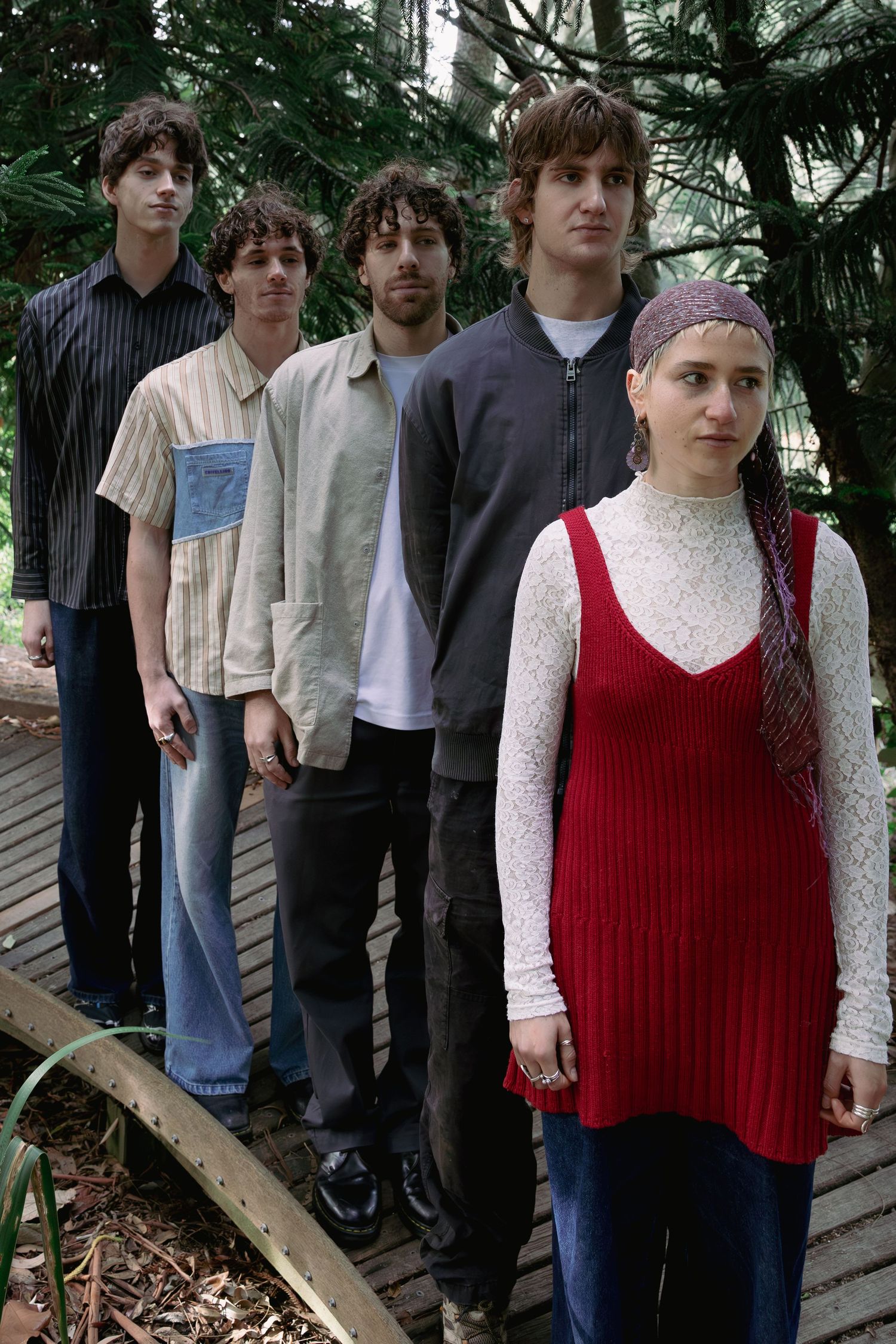 Five young adults posing on a wooden bridge in a lush green park, with a woman in red vest leading the group.