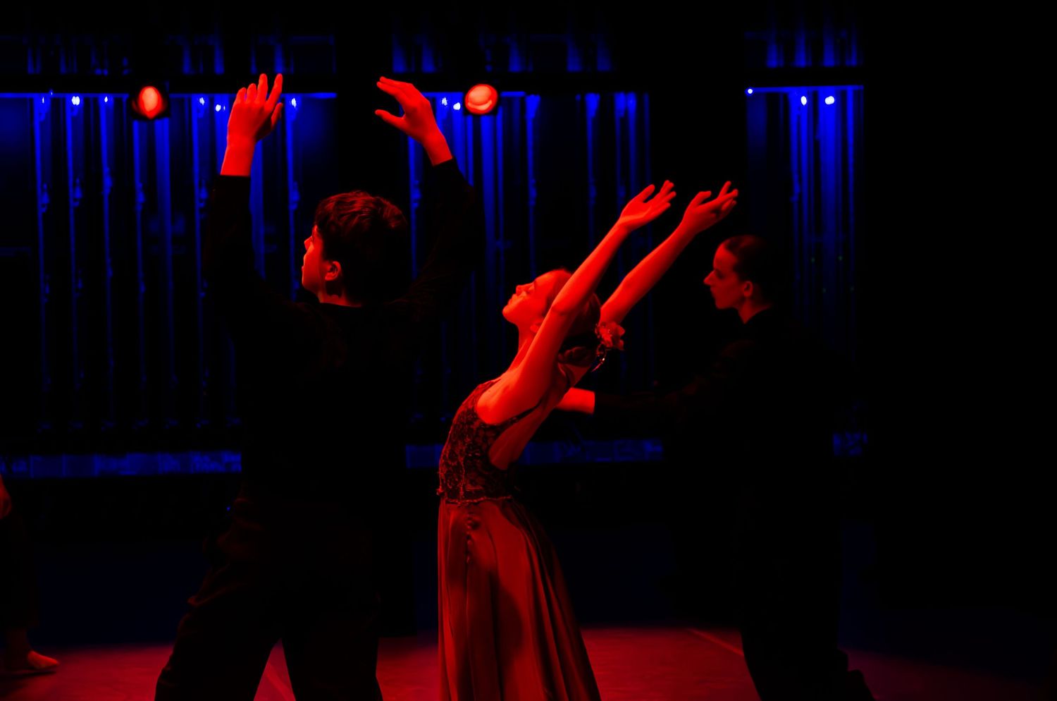 Three dancers perform dramatically on a dimly lit stage with blue and red lighting during a theatrical dance show.