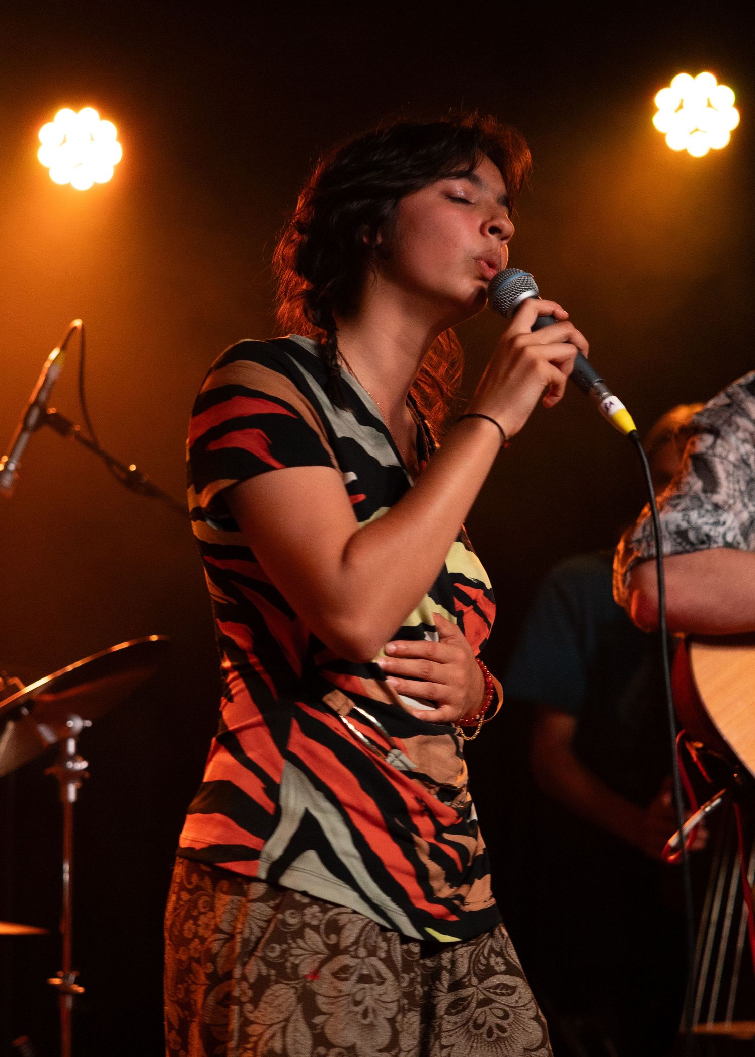 Young woman in zebra-print shirt singing into microphone on stage with warm orange stage lights behind her.