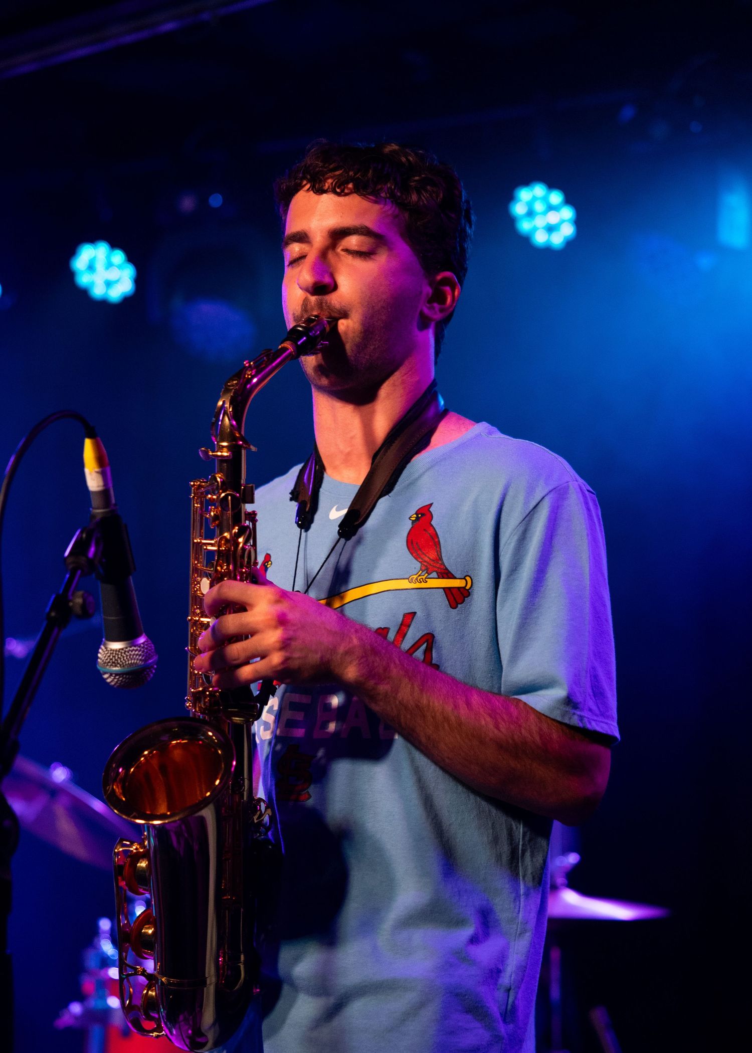 Young man playing alto saxophone on stage under blue and purple lights, wearing a St. Louis Cardinals jersey.