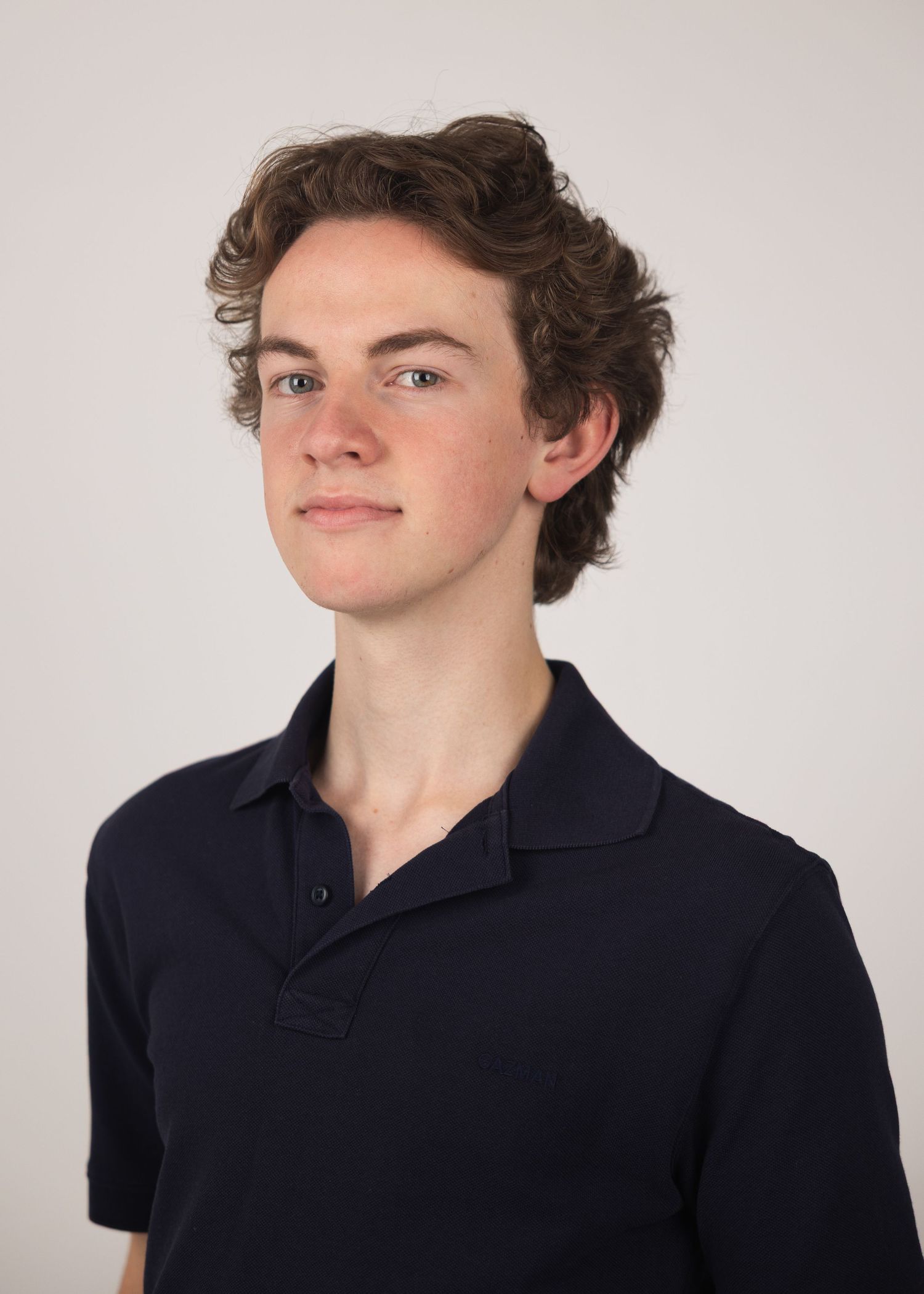Young man with curly brown hair wearing a navy polo shirt poses for a professional headshot portrait.