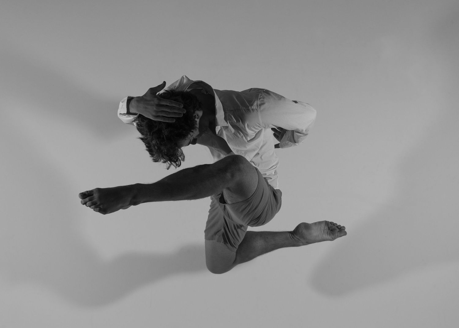 Black and white photo of a dancer mid-leap, bending forward with one leg raised, wearing casual attire.