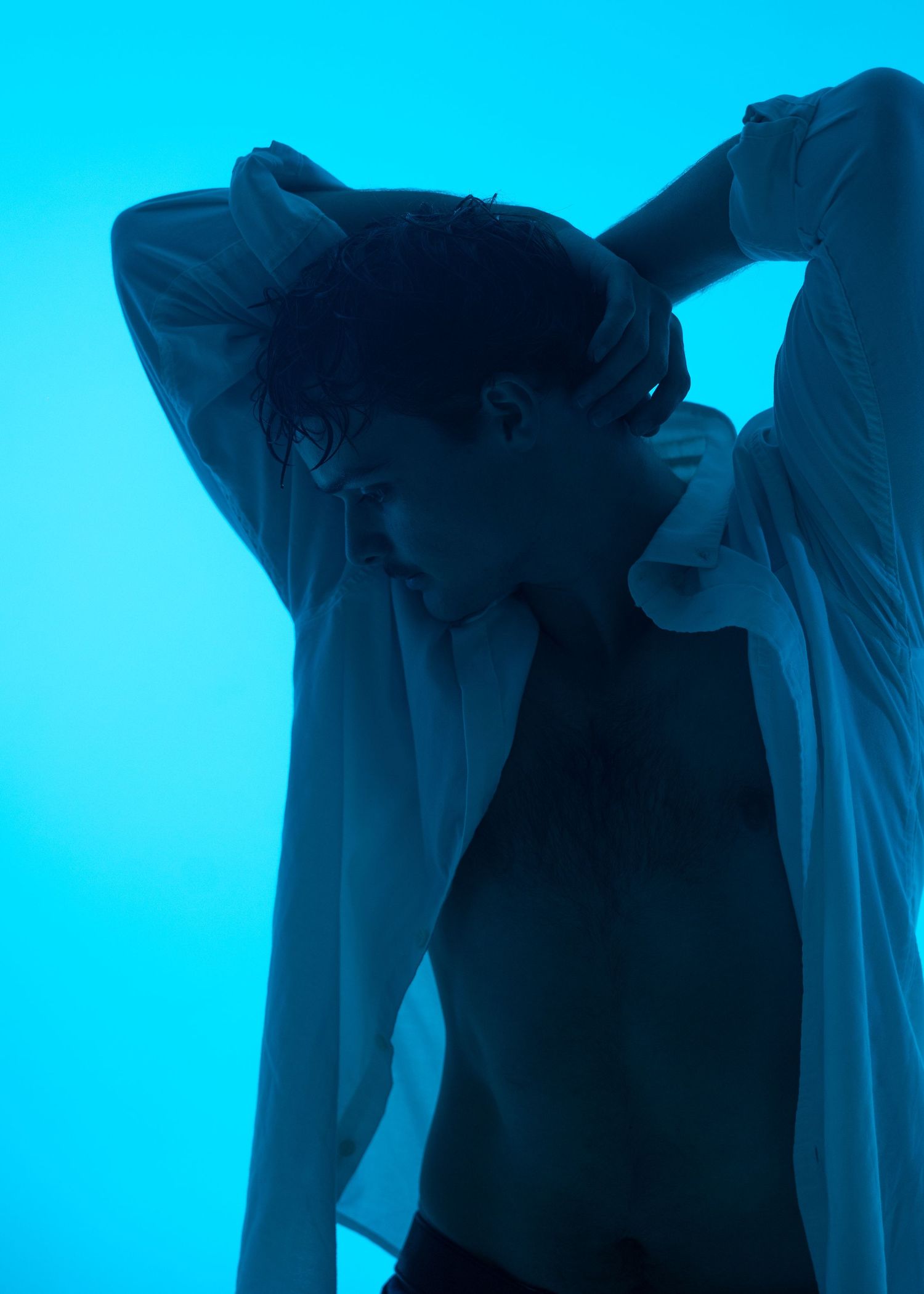 Artistic blue-lit portrait of a person in an open white shirt with hands behind their head.