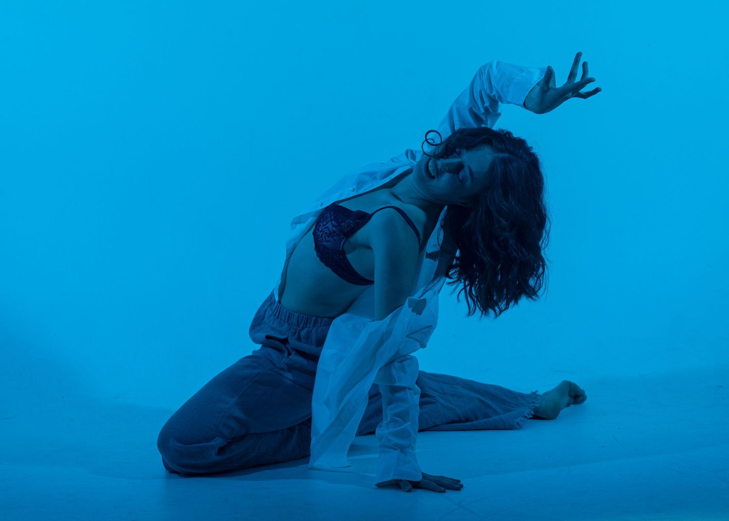 A dancer kneels dramatically on the floor under blue lighting, reaching upward with expressive arms and flowing white fabric.
