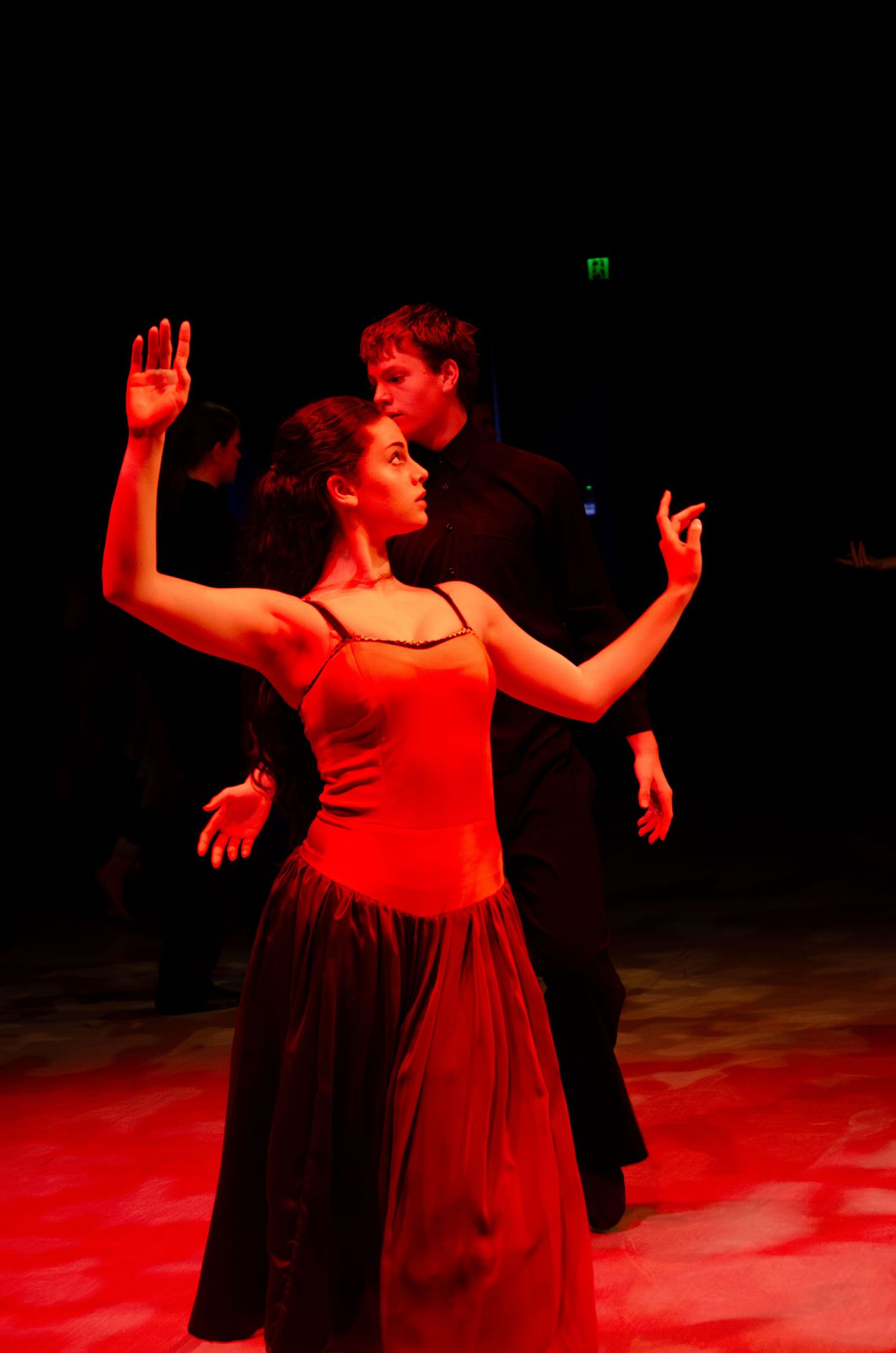 A woman in a red dress dances gracefully under red stage lighting while a man in black watches her performance.