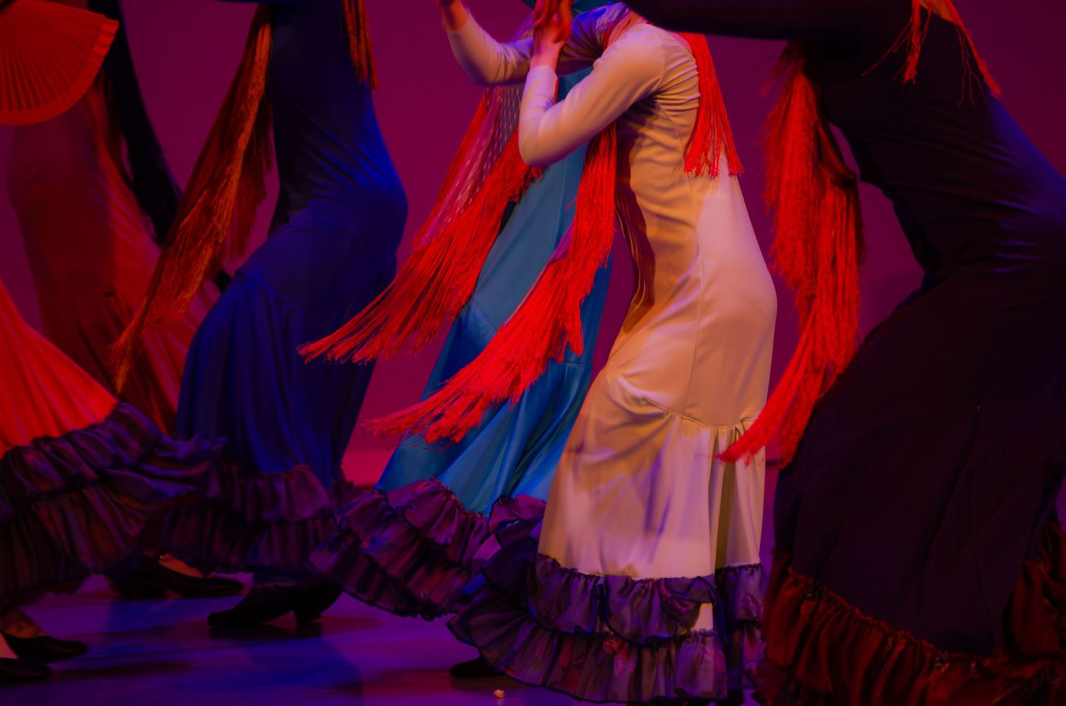 Performers in colorful costumes dance during a theatrical stage production with dramatic red and blue lighting.