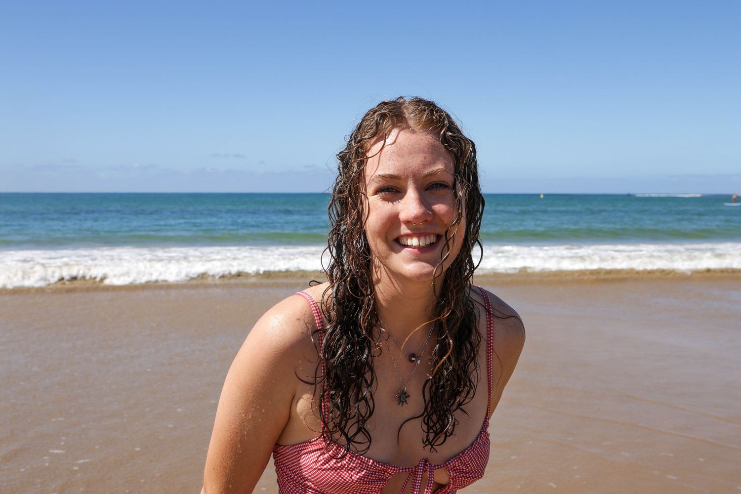 Smiling young woman with wet curly hair wearing a pink bikini top standing on a sunny sandy beach with ocean waves.