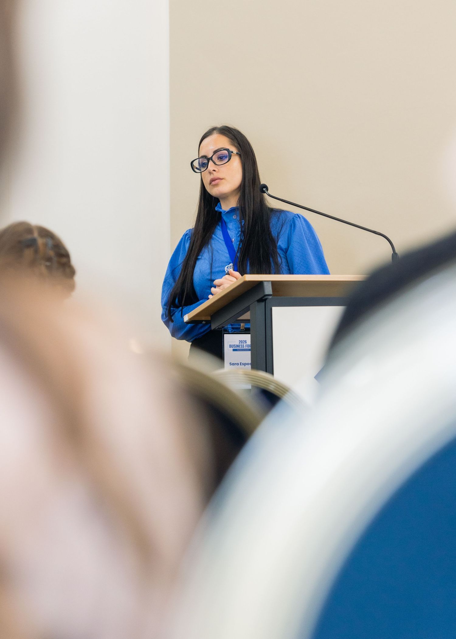 Woman in blue blazer and glasses speaks at podium during professional conference presentation.