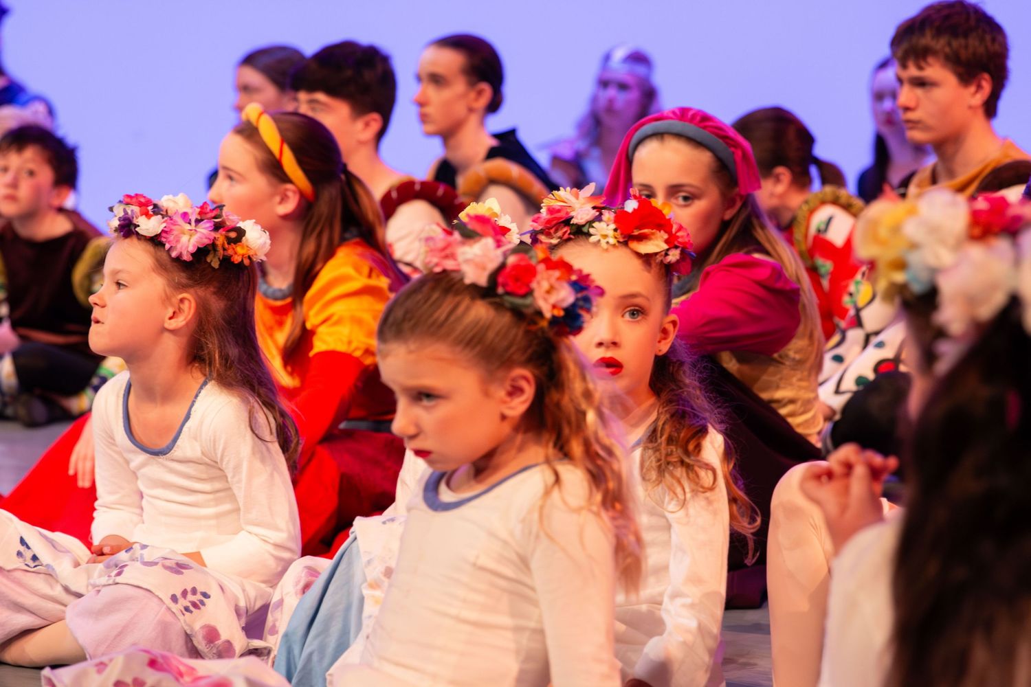 Children in colorful floral costumes sit together during a festive school performance or cultural dance recital.