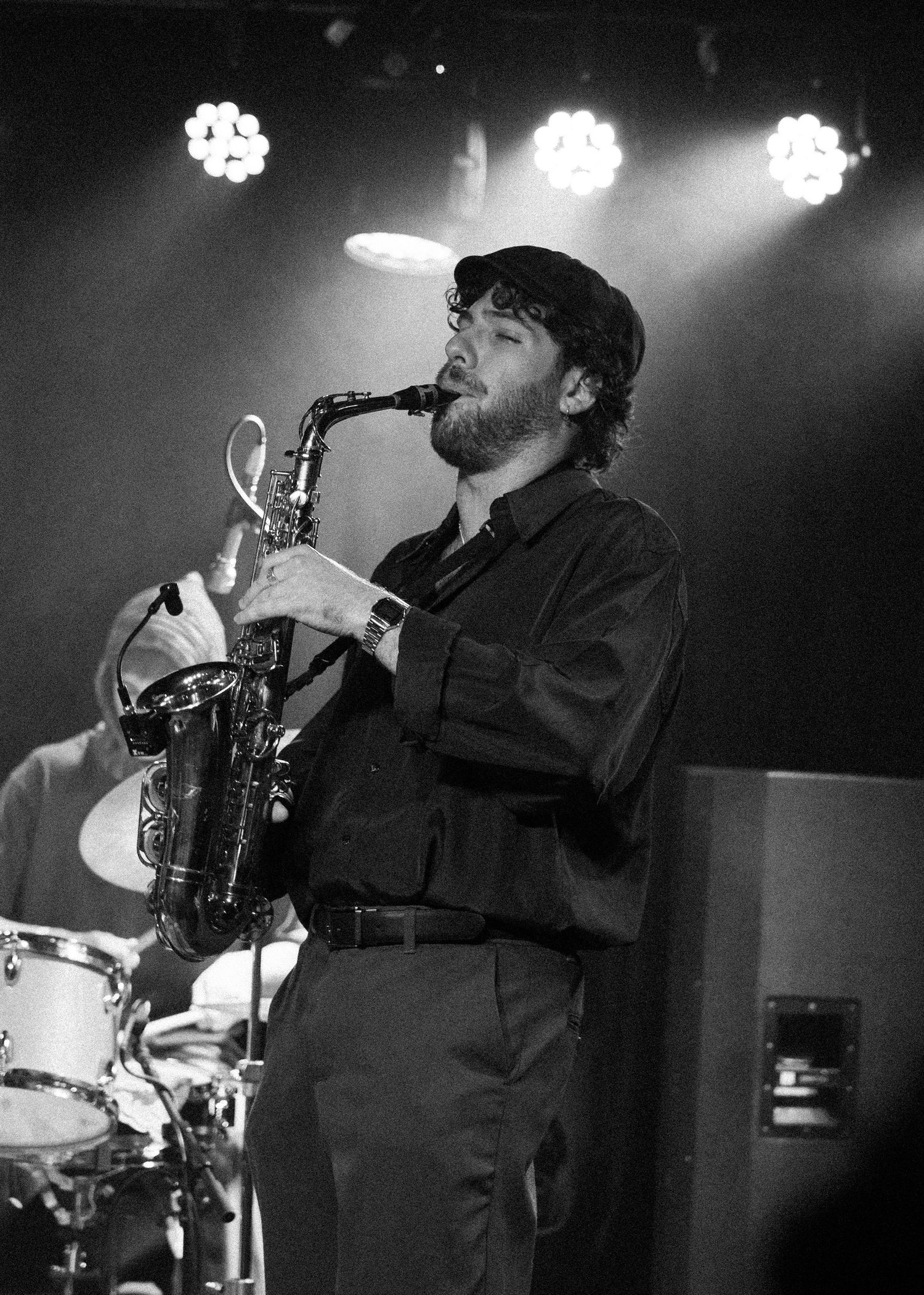 Black and white photo of a bearded man in a cap playing saxophone on stage under bright spotlights.