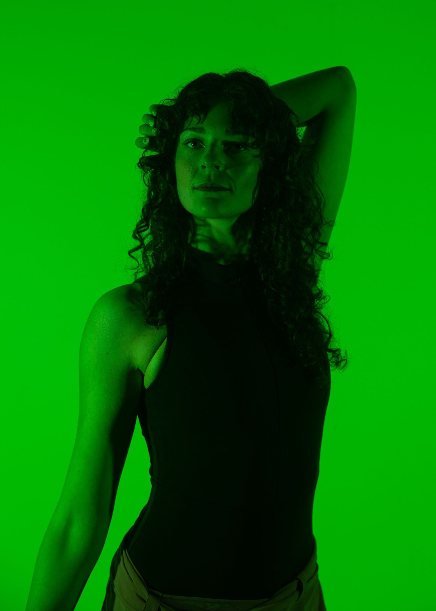 Woman with curly hair posing dramatically under vibrant green lighting in a black sleeveless top.