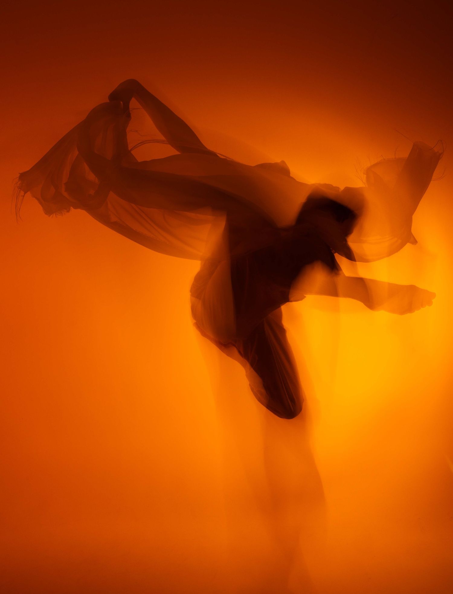 Silhouetted figure dancing with flowing fabric against a warm orange backlight creates a dramatic artistic effect.