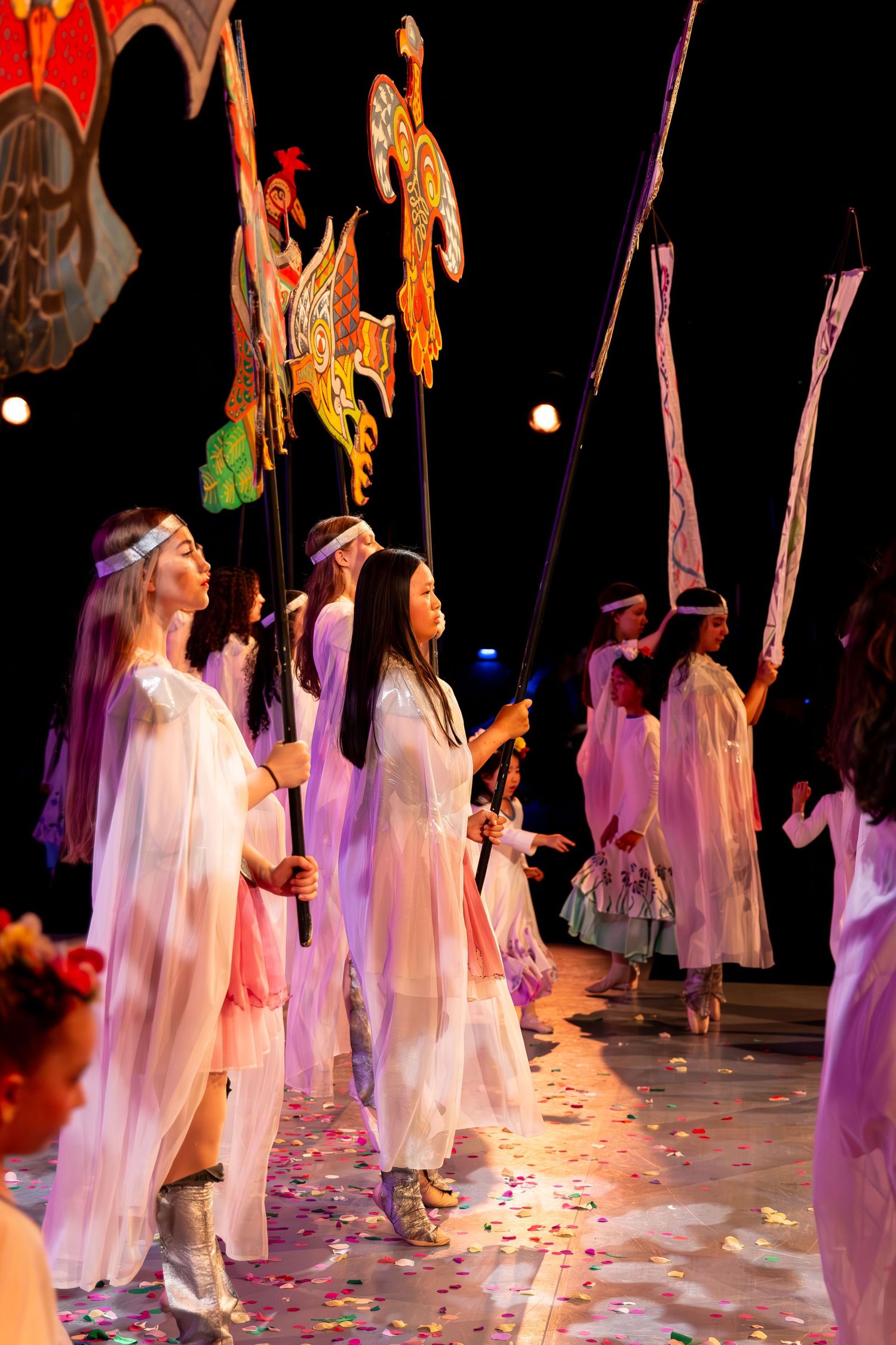 Performers in white robes carry decorative banners on stage amid colorful confetti during a theatrical parade performance.