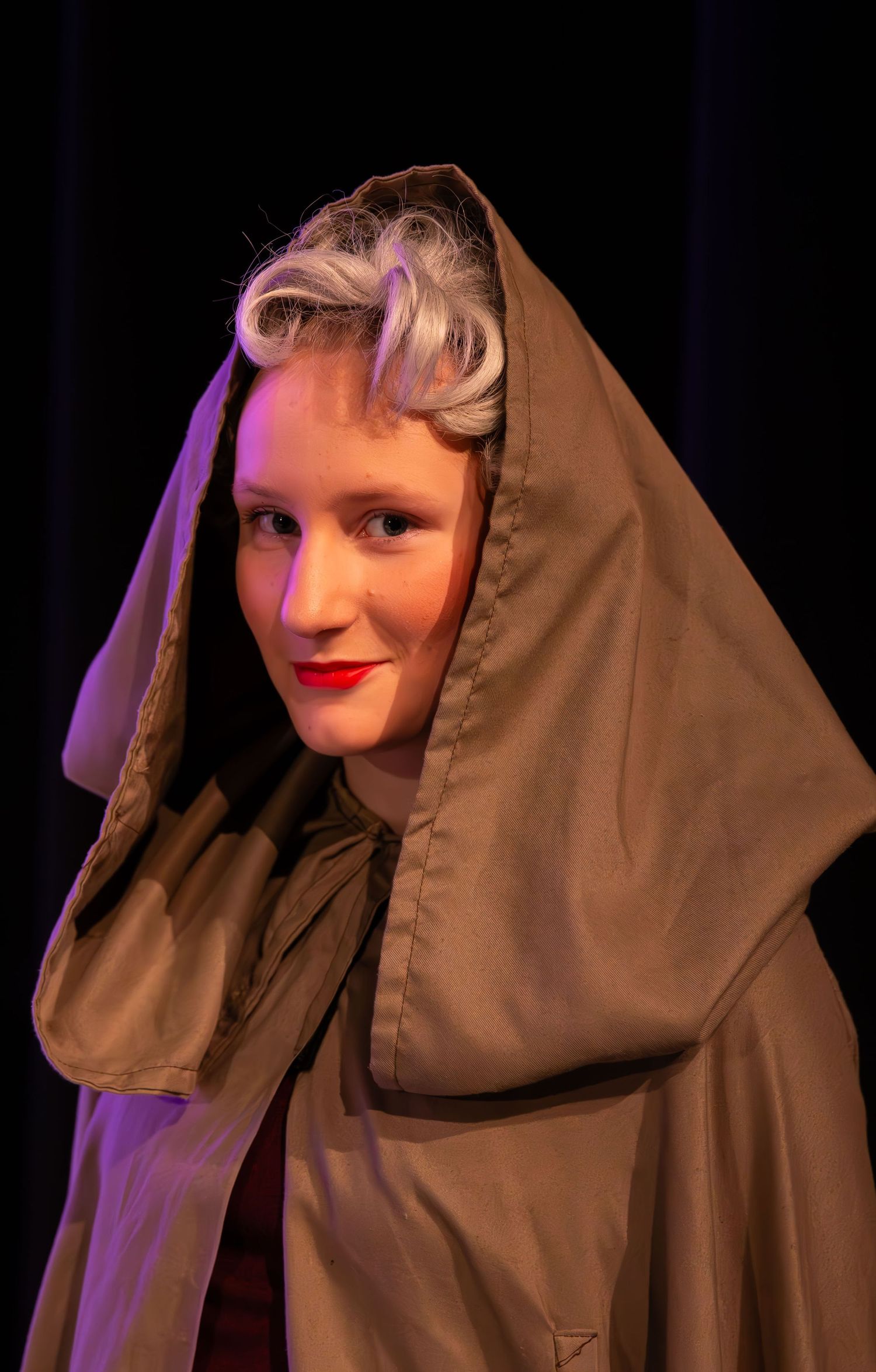 A performer in a hooded olive cloak with styled gray hair and red lips poses on stage.