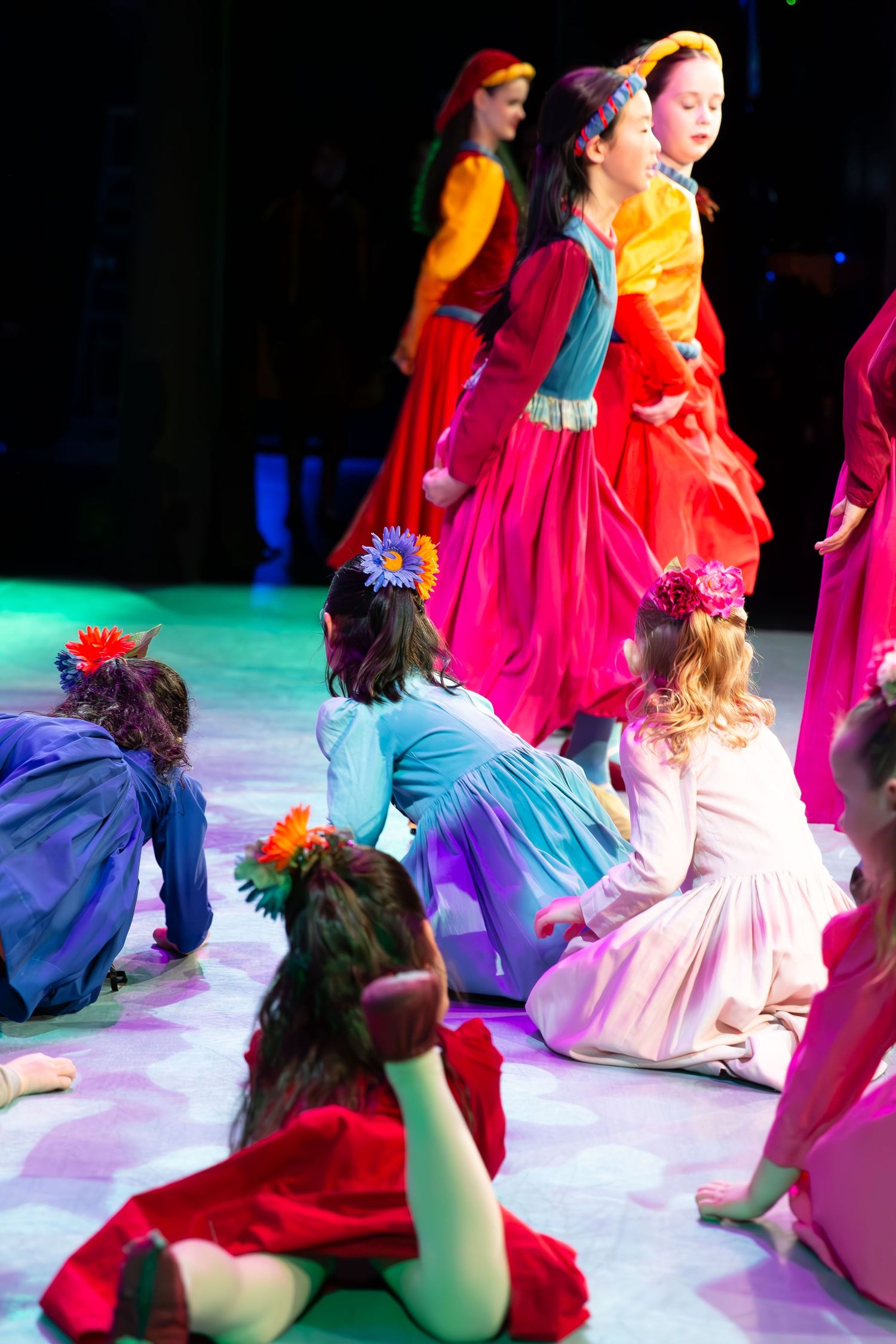 Children in colorful folk costumes perform a dance recital on stage, wearing flower crowns under bright theatrical lighting.