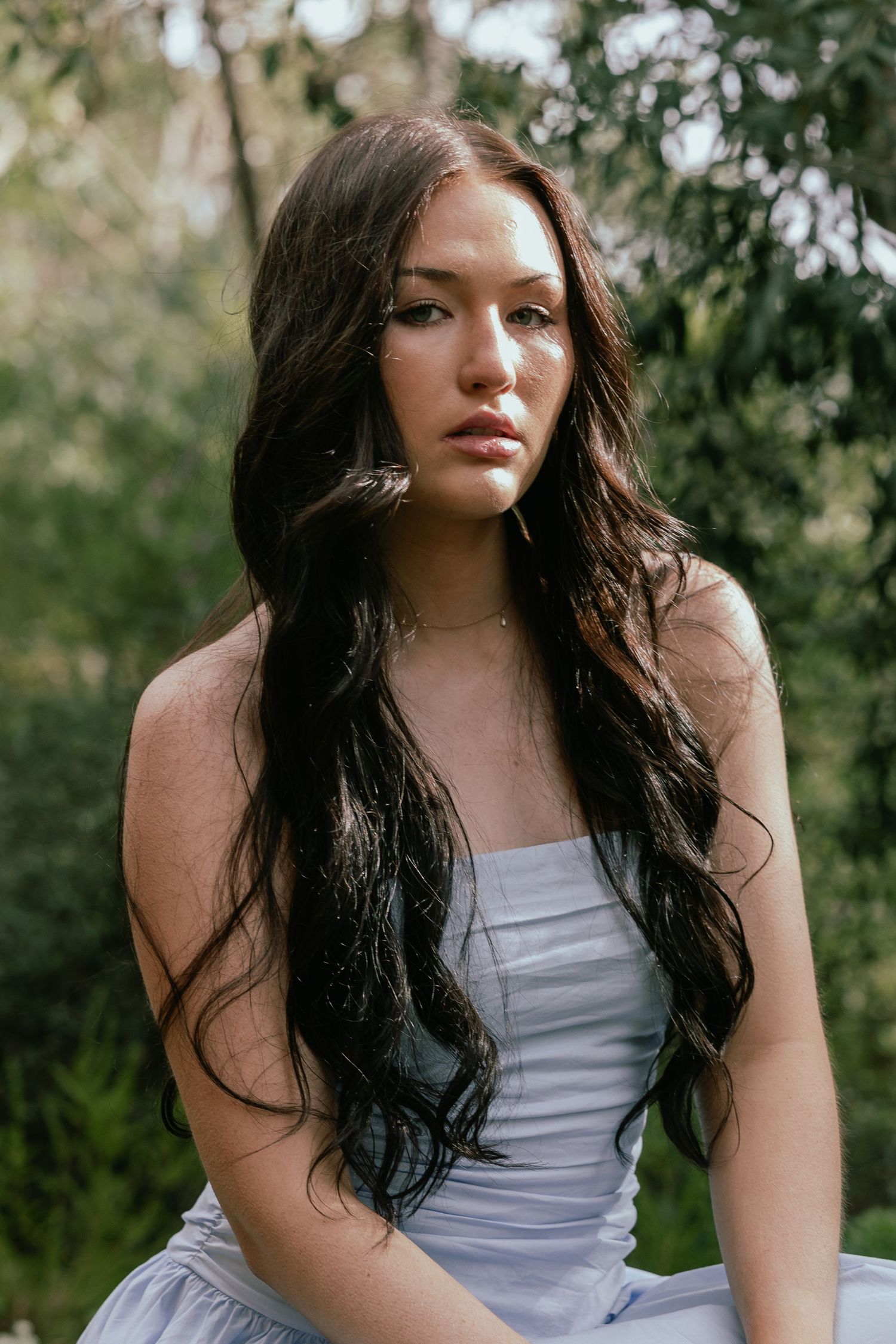 Young woman with long dark wavy hair wearing a light blue dress, sitting outdoors surrounded by lush green trees.