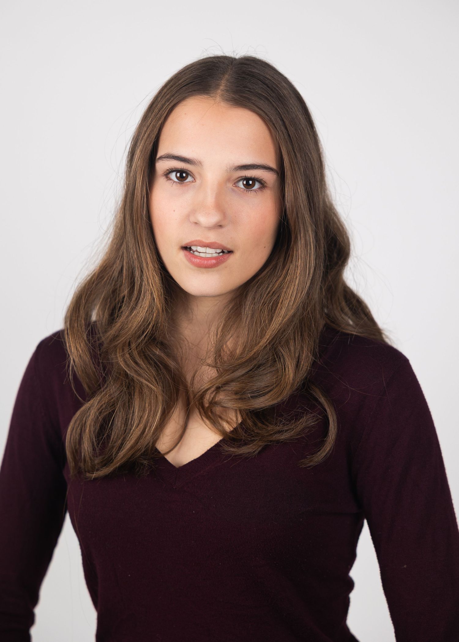 Young woman with long brown wavy hair wearing a burgundy v-neck sweater poses for a professional headshot.