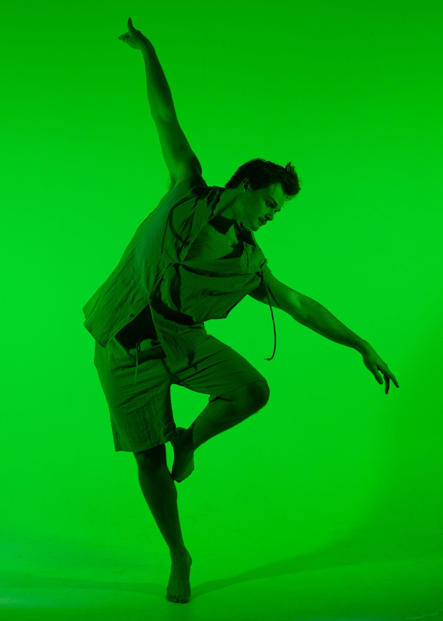 Male dancer leaps gracefully in hooded outfit under vivid green studio lighting.