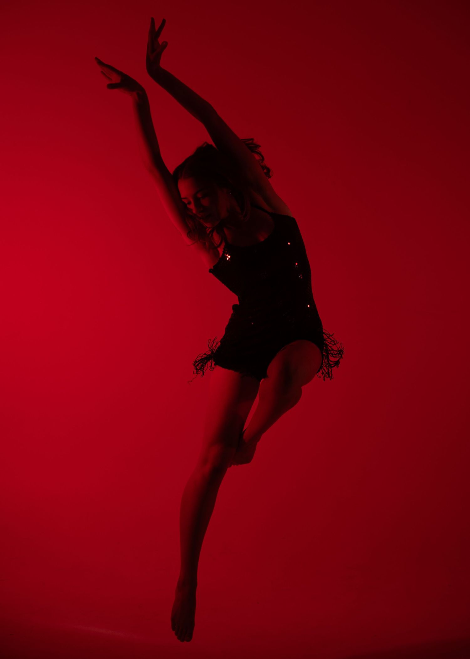 A dancer leaps gracefully against a vivid red background, wearing a black sequined outfit with arms raised elegantly.