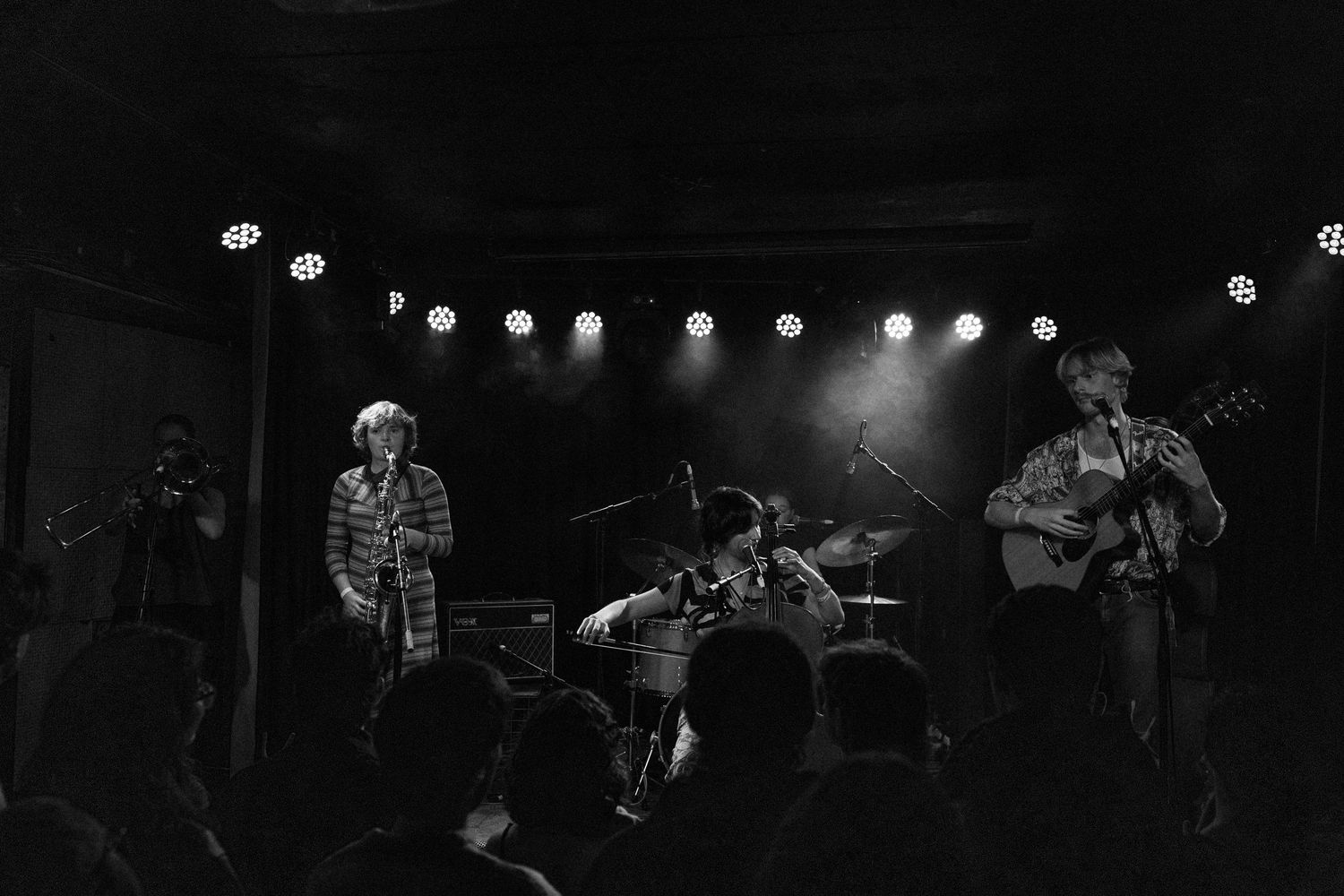 Black and white photo of a live band performing on a dimly lit stage with bright overhead lights and an audience.