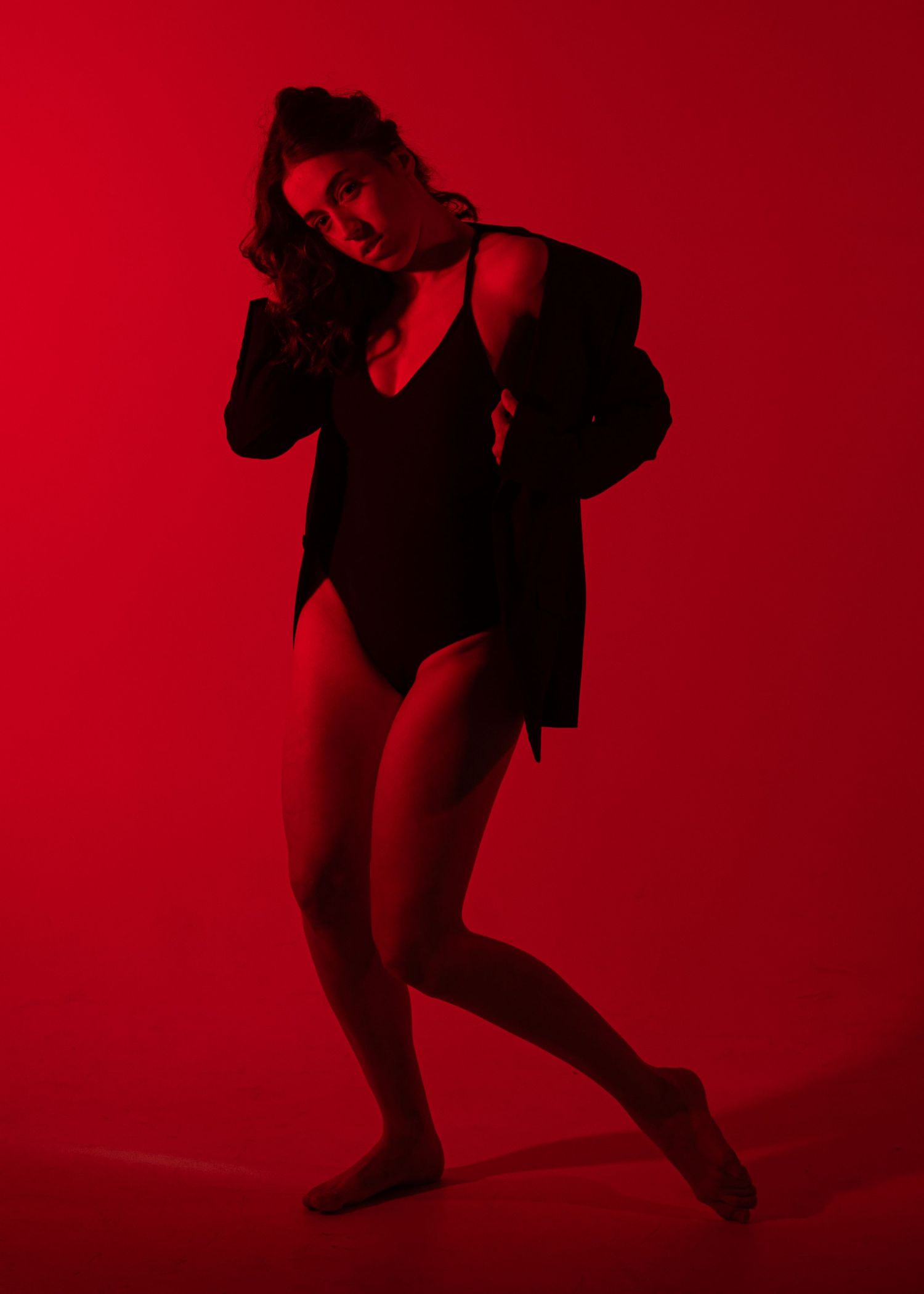 Woman posing dramatically in black bodysuit and jacket against a bold red background.