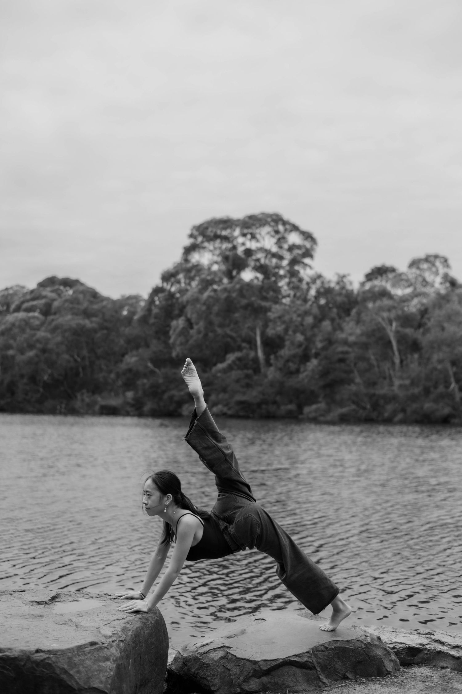 Woman performing a yoga pose on rocks by a serene lake, surrounded by lush trees, in black and white.