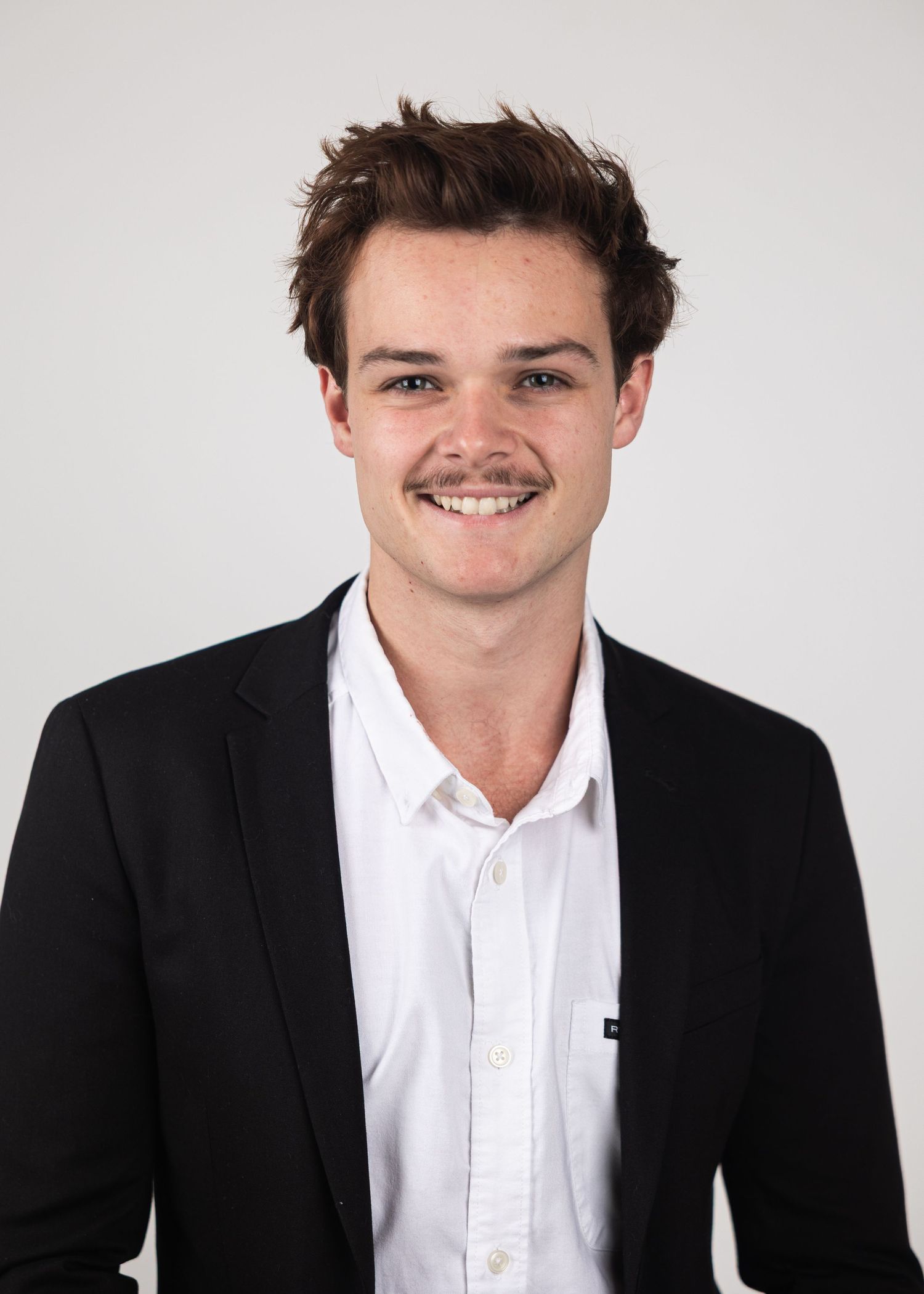 Smiling young man in black blazer and white shirt poses for professional headshot.