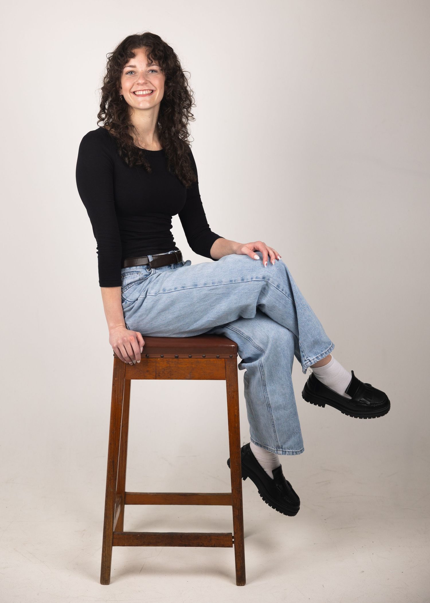 Woman with curly dark hair sitting on a wooden stool, wearing black top and light blue jeans.