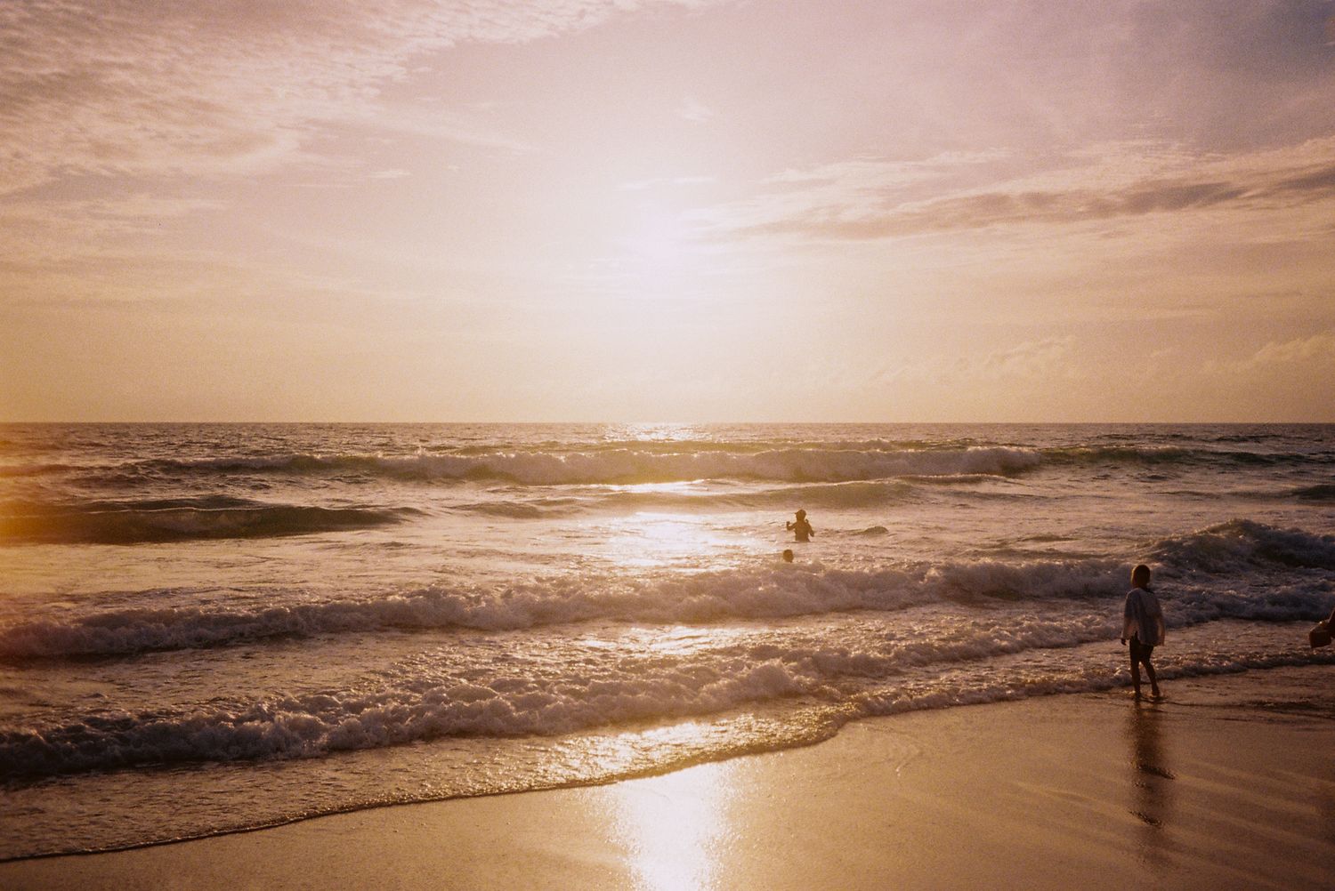 Two people enjoy a stunning golden sunset on a sandy beach with gentle waves rolling in.