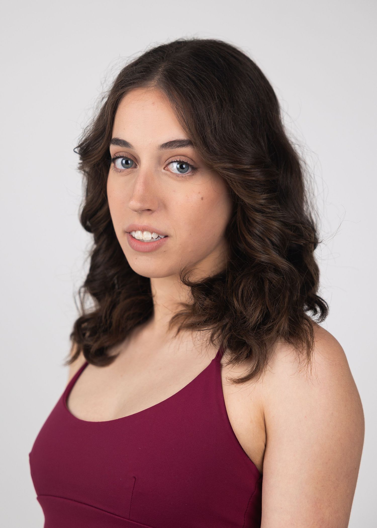 Young woman with wavy brown hair and blue eyes wearing a burgundy tank top against a white background.