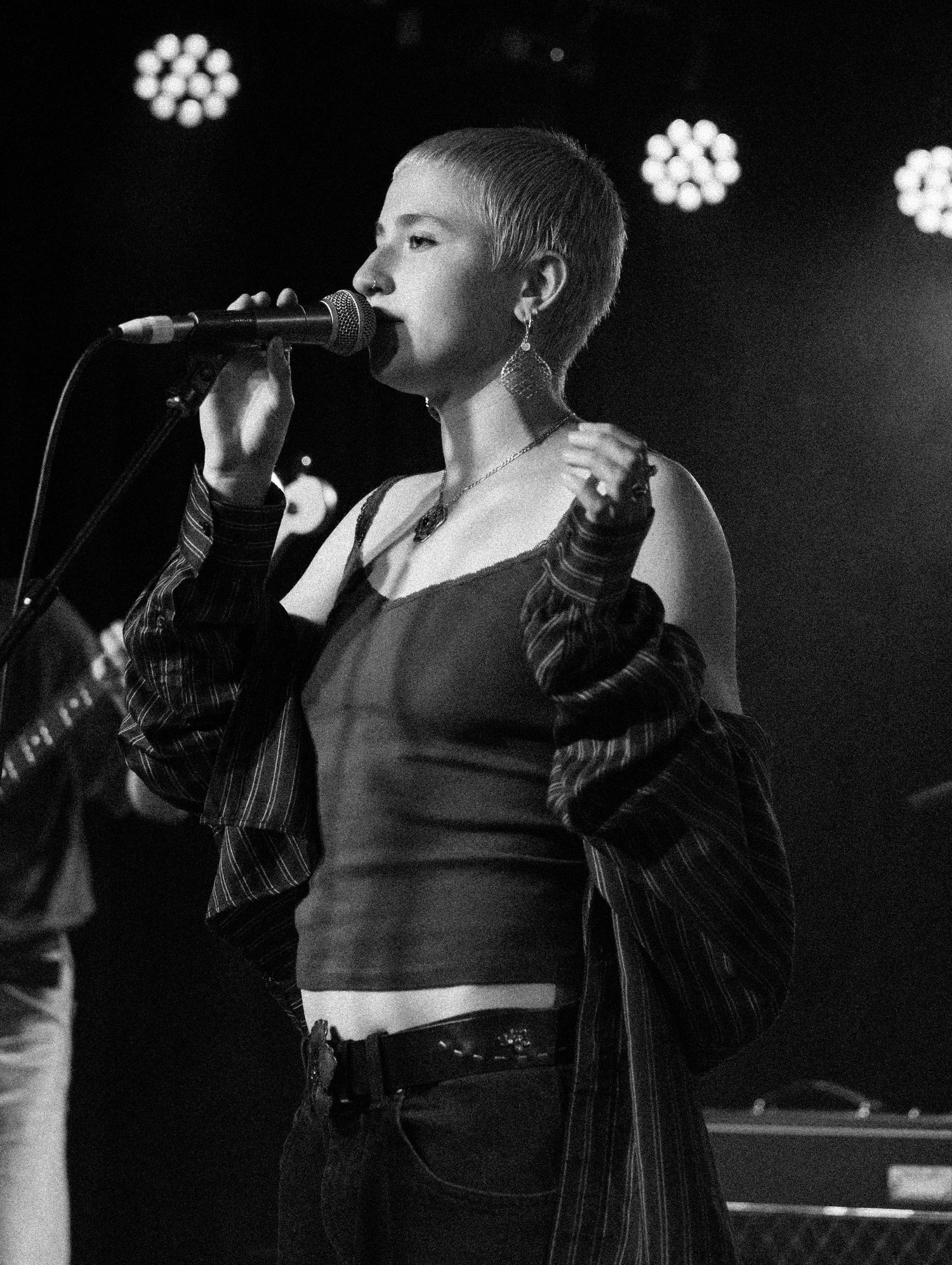 Female singer with shaved head performing on stage with microphone under stage lights.