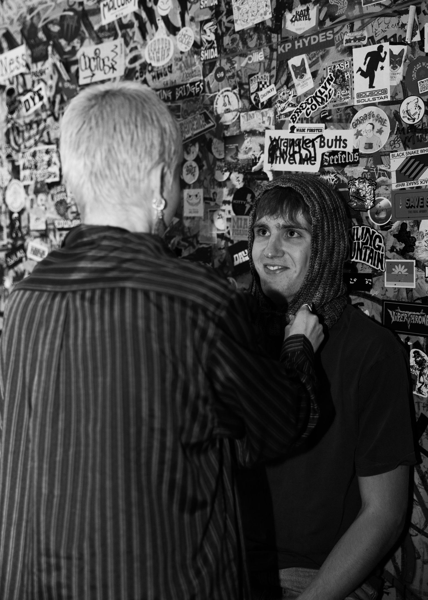 Black and white photo of two people talking in front of a wall covered in stickers and posters.